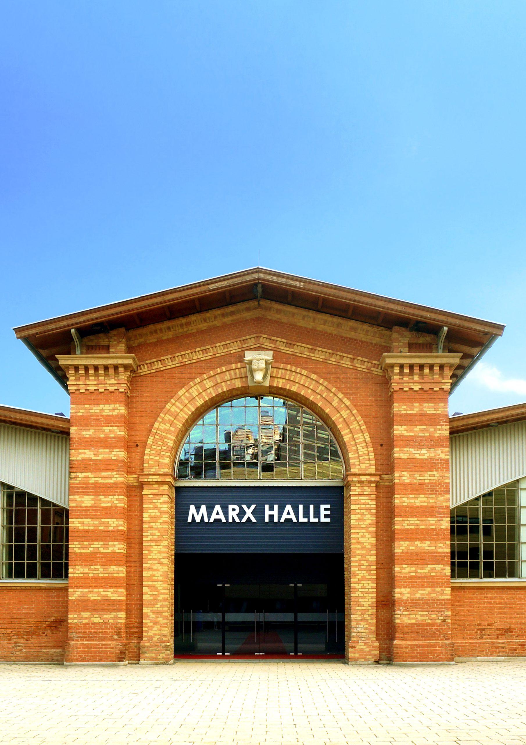 Ein rotes Backsteingebäude mit einem großen gewölbten Eingang, über dessen Glastüren ein Schild mit der Aufschrift MARX HALLE prangt, mit blauem Himmel über dem Kopf und Spiegelungen in den Fenstern.