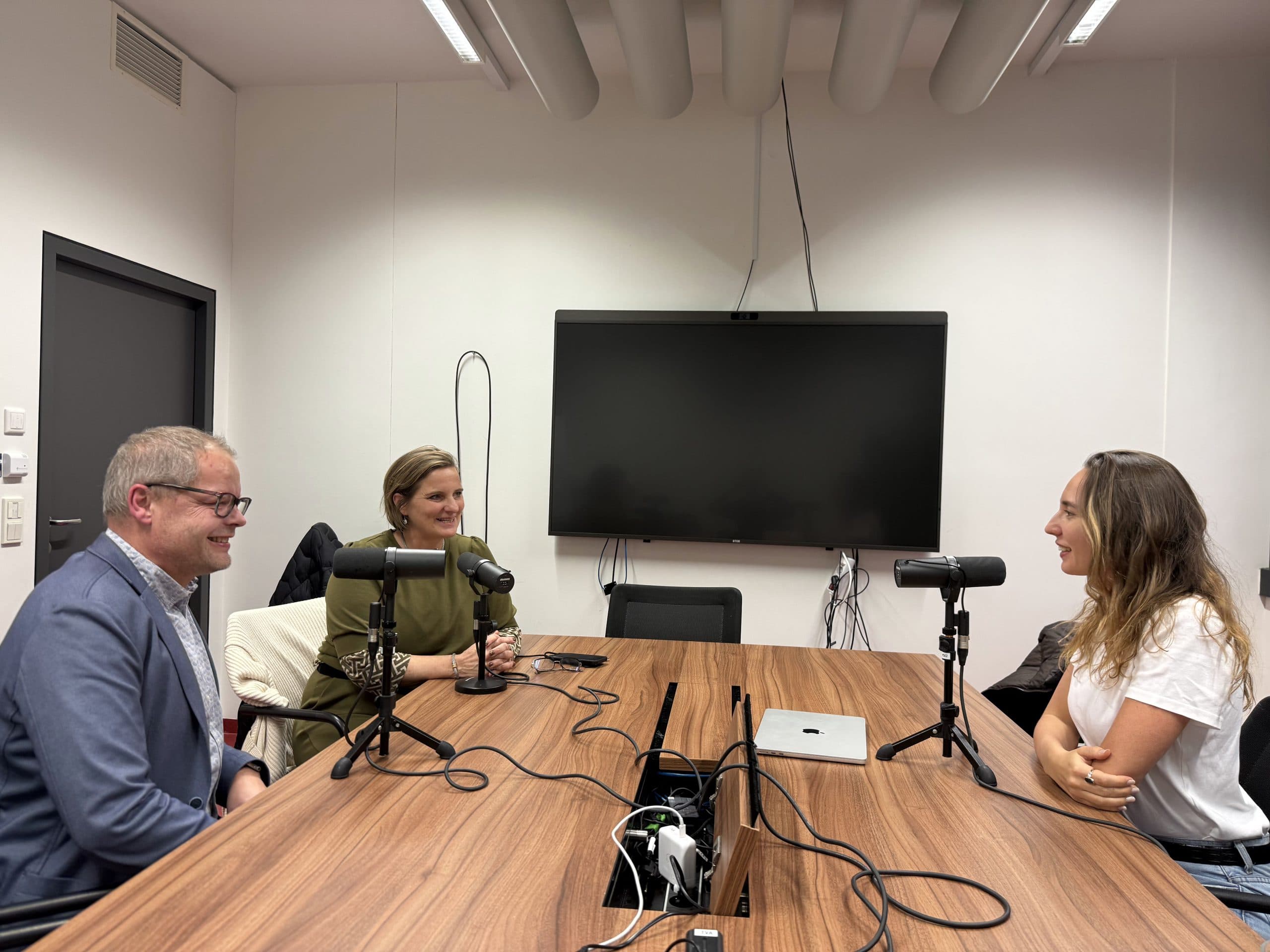 Drei Personen sitzen um einen Tisch mit Mikrofonen vor sich und nehmen in einem modernen Büroraum einen Podcast auf. An der Wand hinter ihnen ist ein großer Monitor angebracht, und auf dem Tisch sind Kabel zu sehen.