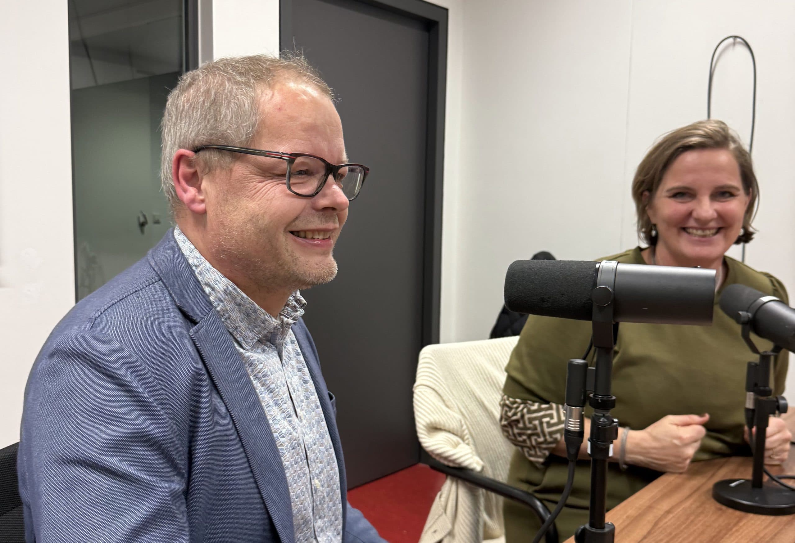 Ein Mann und eine Frau sitzen an einem Tisch mit Mikrofonen und lächeln, als ob sie einen Podcast aufnehmen oder ein Gespräch in einer Studioumgebung führen würden.