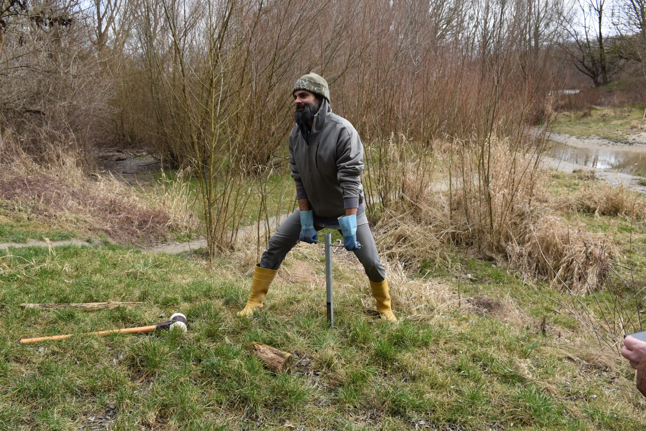 Eine Person in Outdoor-Kleidung, Handschuhen und Stiefeln steht im Gras und gräbt mit einem Metallwerkzeug in den Boden. Im Hintergrund sind blattlose Sträucher zu sehen und in der Nähe liegen Werkzeuge.
