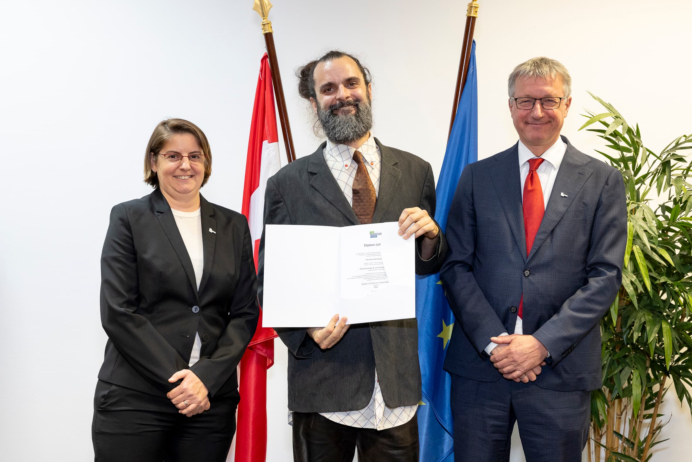 Drei Personen in Geschäftskleidung stehen im Haus und lächeln. Die Person in der Mitte hält eine Urkunde in der Hand. Hinter ihnen sind eine kanadische Flagge, eine EU-Flagge und eine Blattpflanze zu sehen.