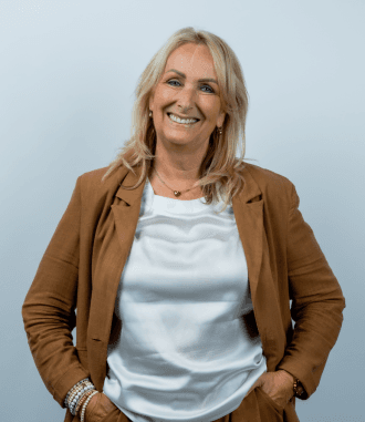 Eine lächelnde Frau mit blondem Haar trägt einen braunen Blazer über einer weißen Bluse und ist mit Armbändern und einer Halskette geschmückt. Sie steht vor einem schlichten hellen Hintergrund und hat die Hände in den Taschen.