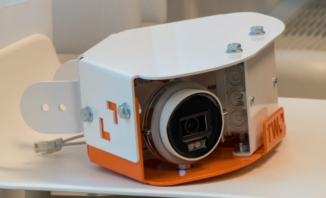 Ein weißes und orangefarbenes elektronisches Gerät mit einer Linse, möglicherweise eine Kamera oder ein Sensor, sitzt auf einem weißen Stuhl in der Nähe eines Fensters mit durchsichtigen Vorhängen und einem Heizkörper im Hintergrund.