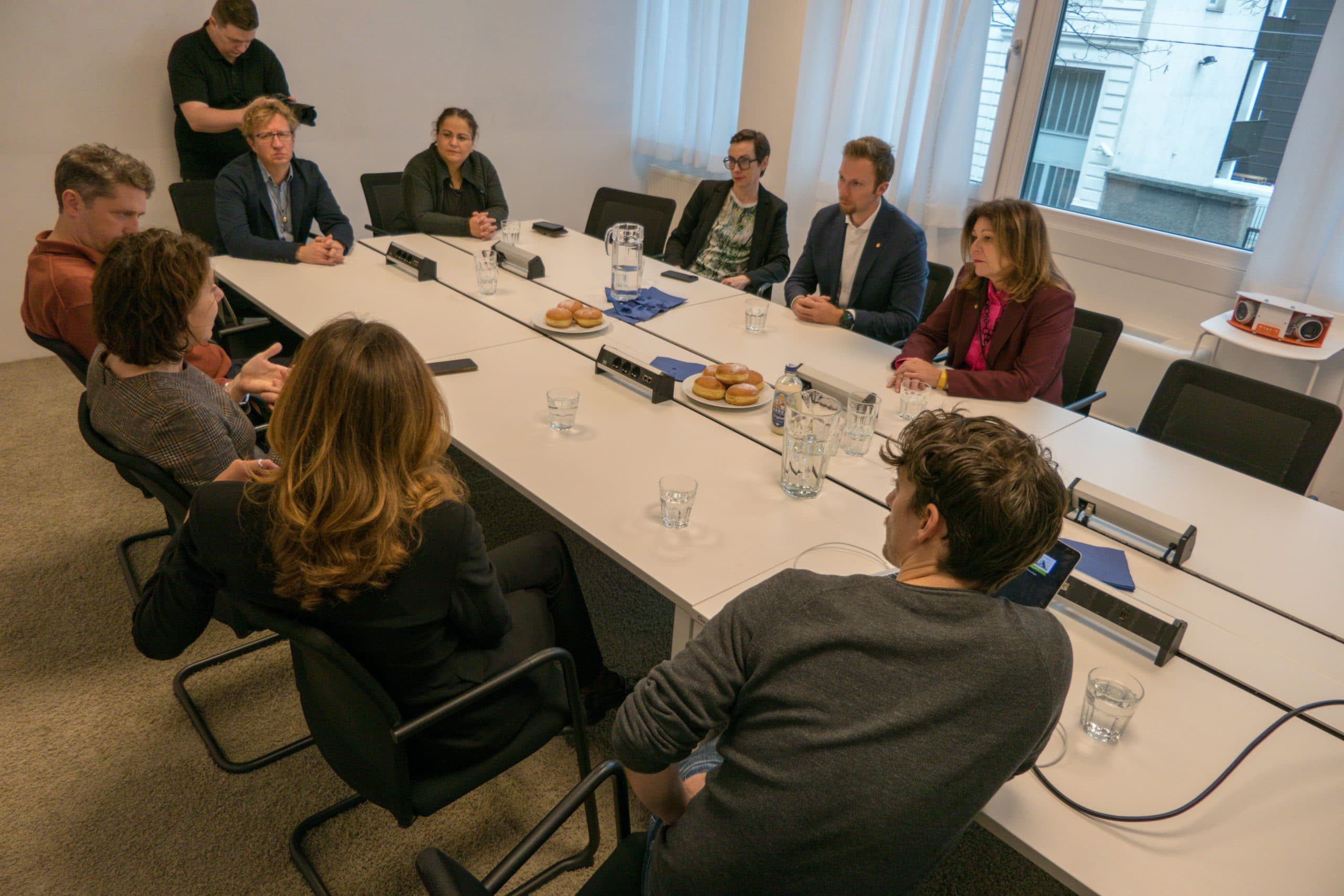 Acht Personen sitzen um einen rechteckigen Konferenztisch in einem Büro und diskutieren. Auf dem Tisch befinden sich Namensschilder, Donuts, Wassergläser und Notizblöcke. Eine Person steht im Hintergrund und filmt die Besprechung.