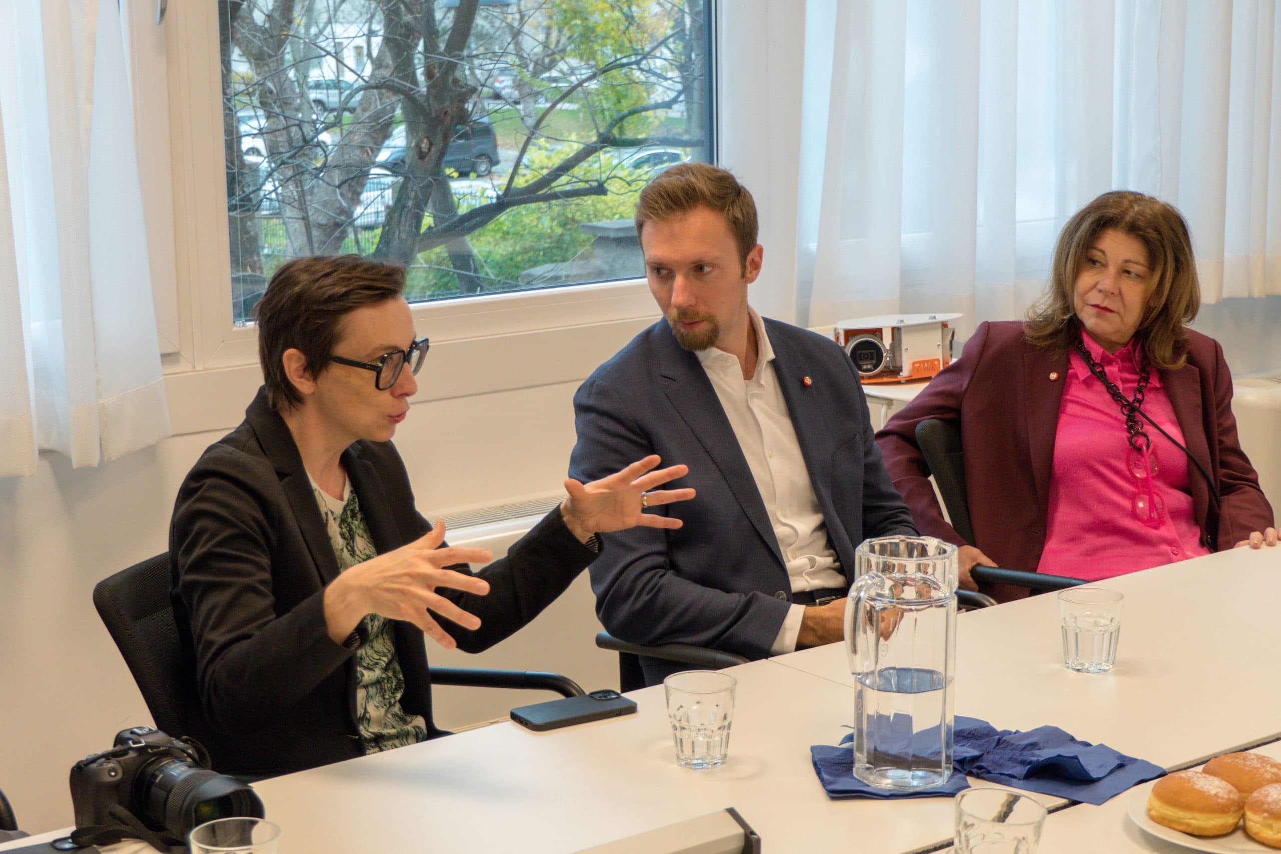 Drei Personen sitzen an einem Konferenztisch und diskutieren. Die Person auf der linken Seite gestikuliert mit beiden Händen, während die beiden anderen zuhören. Auf dem Tisch stehen ein Glaskrug, Gläser, ein Gebäckstück und eine Kamera. Im Hintergrund ist ein Fenster zu sehen.