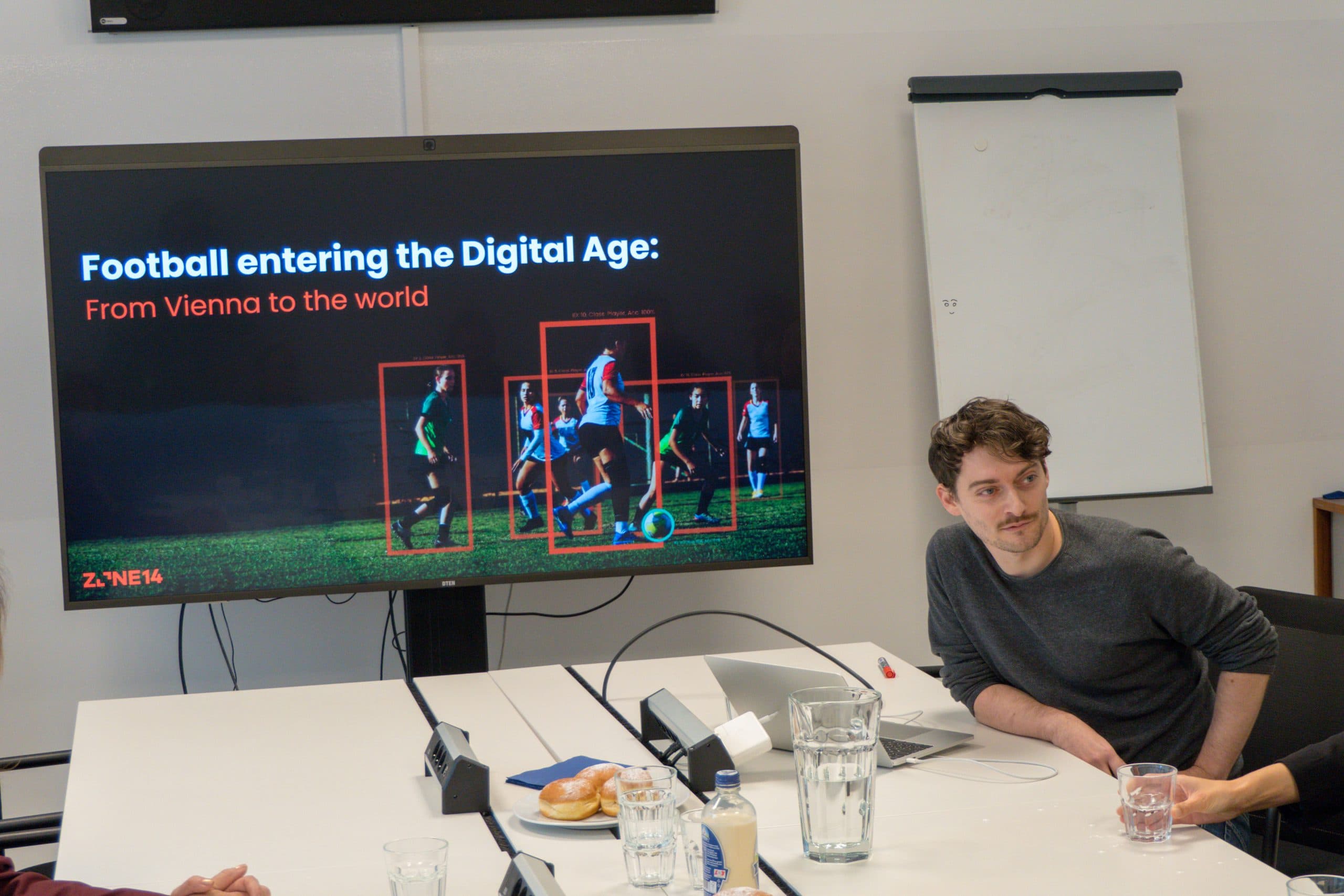 Ein Mann sitzt an einem Konferenztisch neben einem großen Bildschirm, auf dem eine Präsentation mit dem Titel 'Fußball im digitalen Zeitalter: Von Wien in die Welt', mit einem Bild von Fußballspielern auf dem Feld.