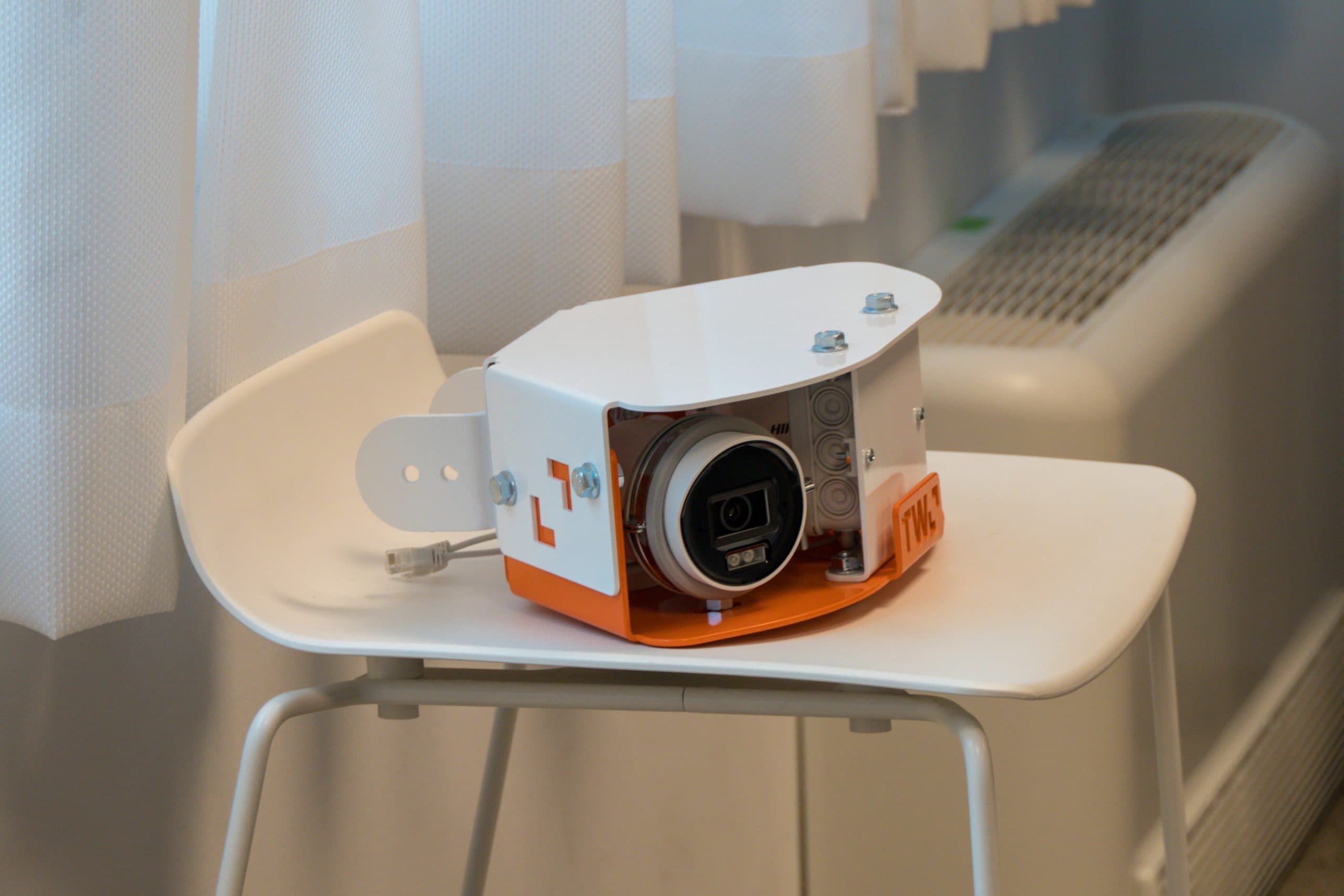Eine weiße und orangefarbene Überwachungskamera ist auf einem weißen Stuhl neben einem Fenster mit weißen Vorhängen in der Nähe eines weißen Heizkörpers angebracht.