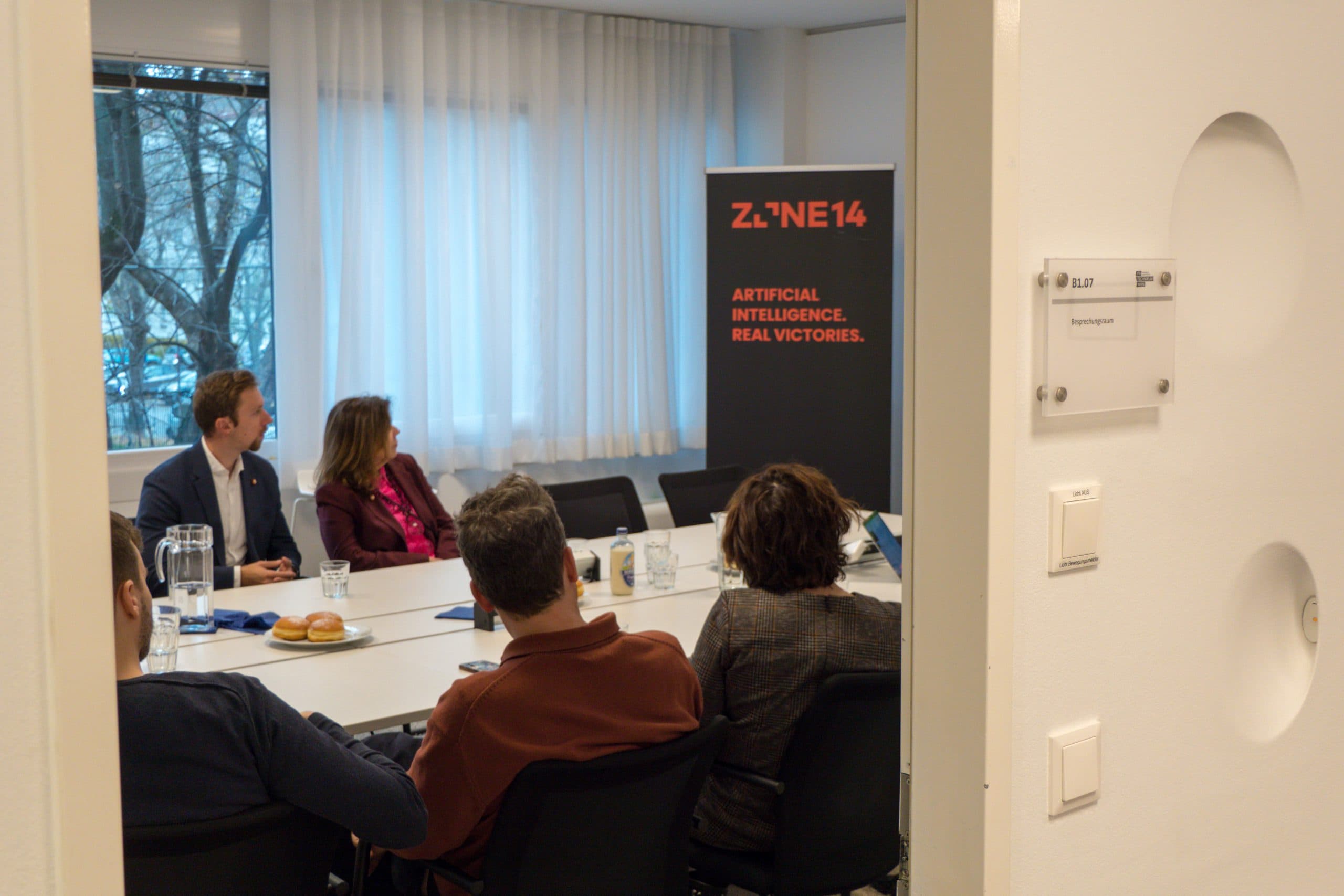 Vier Personen sitzen um einen Konferenztisch in einem Besprechungsraum und blicken auf ein Präsentationsbanner mit der Aufschrift 'ZONE14: Artificial Intelligence. Echte Siege'. Der Raum hat große Fenster mit weißen Vorhängen.
