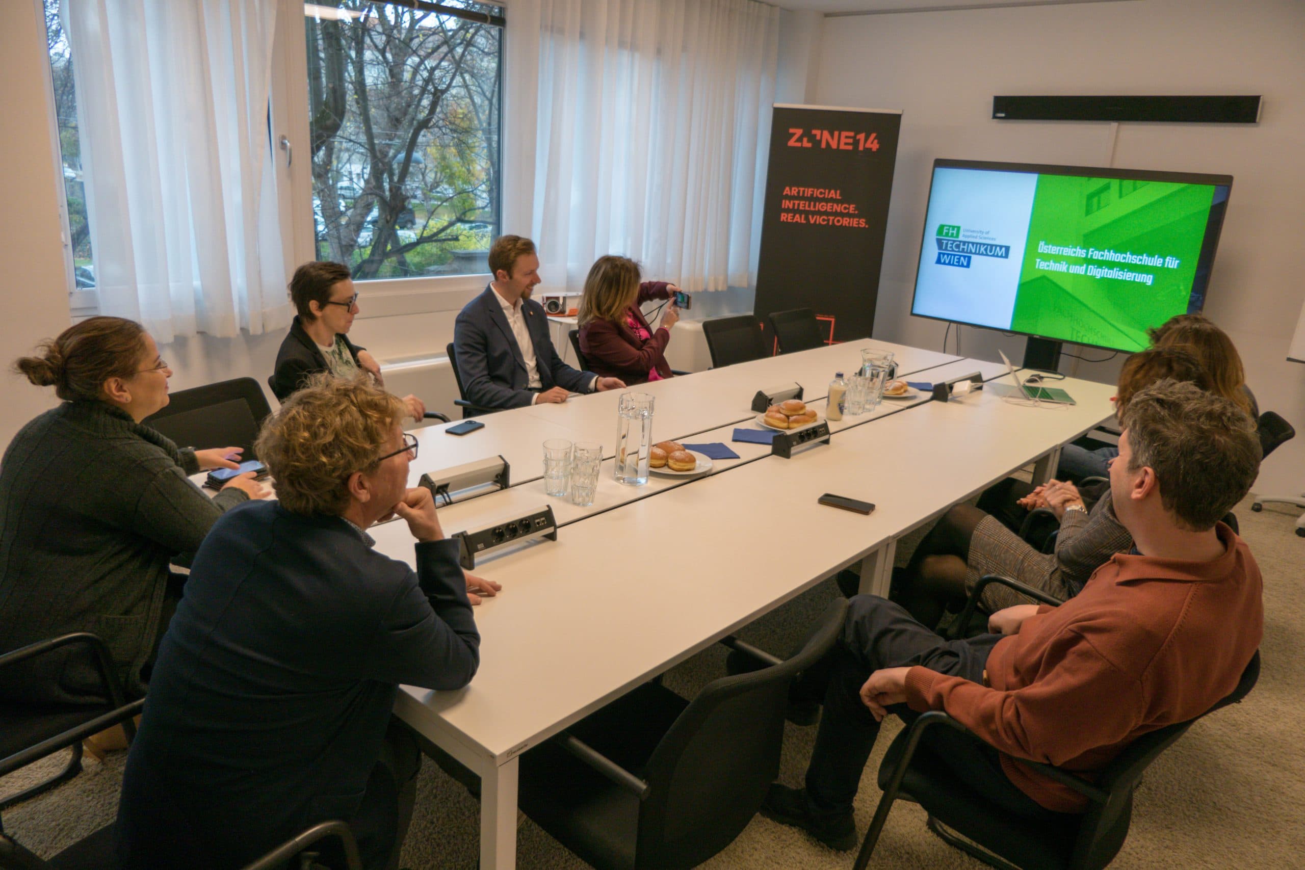 Sieben Personen sitzen um einen Konferenztisch und sehen sich eine Präsentation auf einem großen Bildschirm an, auf dem der Text Kommission Gesellschaftliche Teilhabe und Digitalisierung erscheint. Der Raum ist hell mit großen Fenstern.