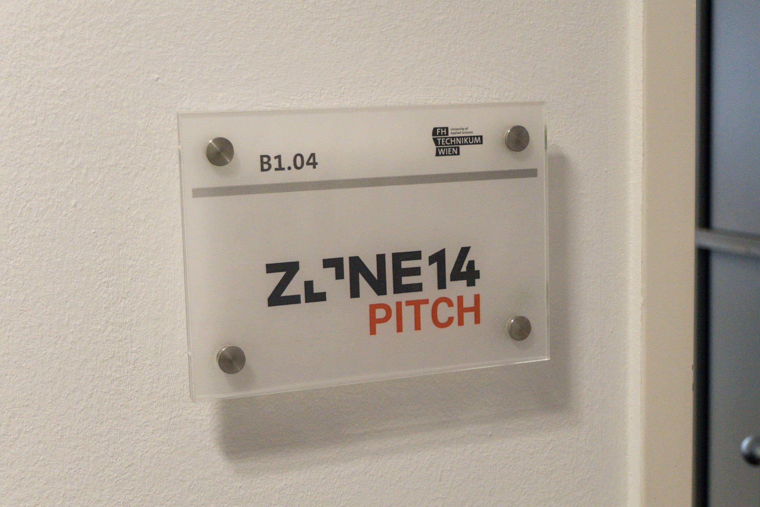 Ein an der Wand angebrachtes Schild trägt oben die Aufschrift B1.04, darunter das Logo der FH Technikum Wien. Darunter in großer Schrift: ZONE14 PITCH, mit PITCH in orange. Das Schild ist mit vier Metallbolzen befestigt.