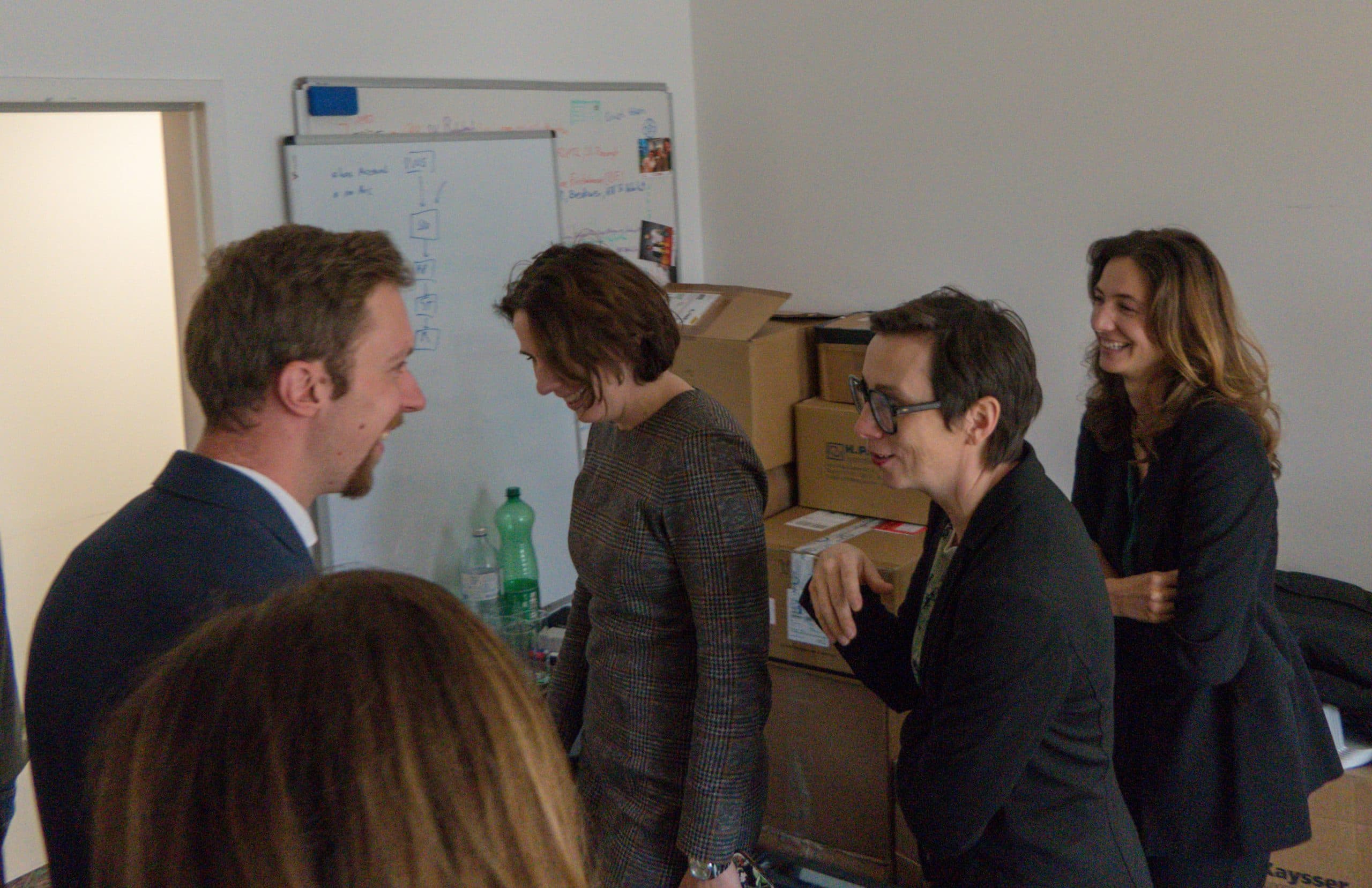 Vier Personen in Geschäftskleidung unterhalten sich und lachen in einem Büro mit Kartons, Flaschen und einem Whiteboard im Hintergrund. Eine Person ist teilweise im Vordergrund zu sehen.