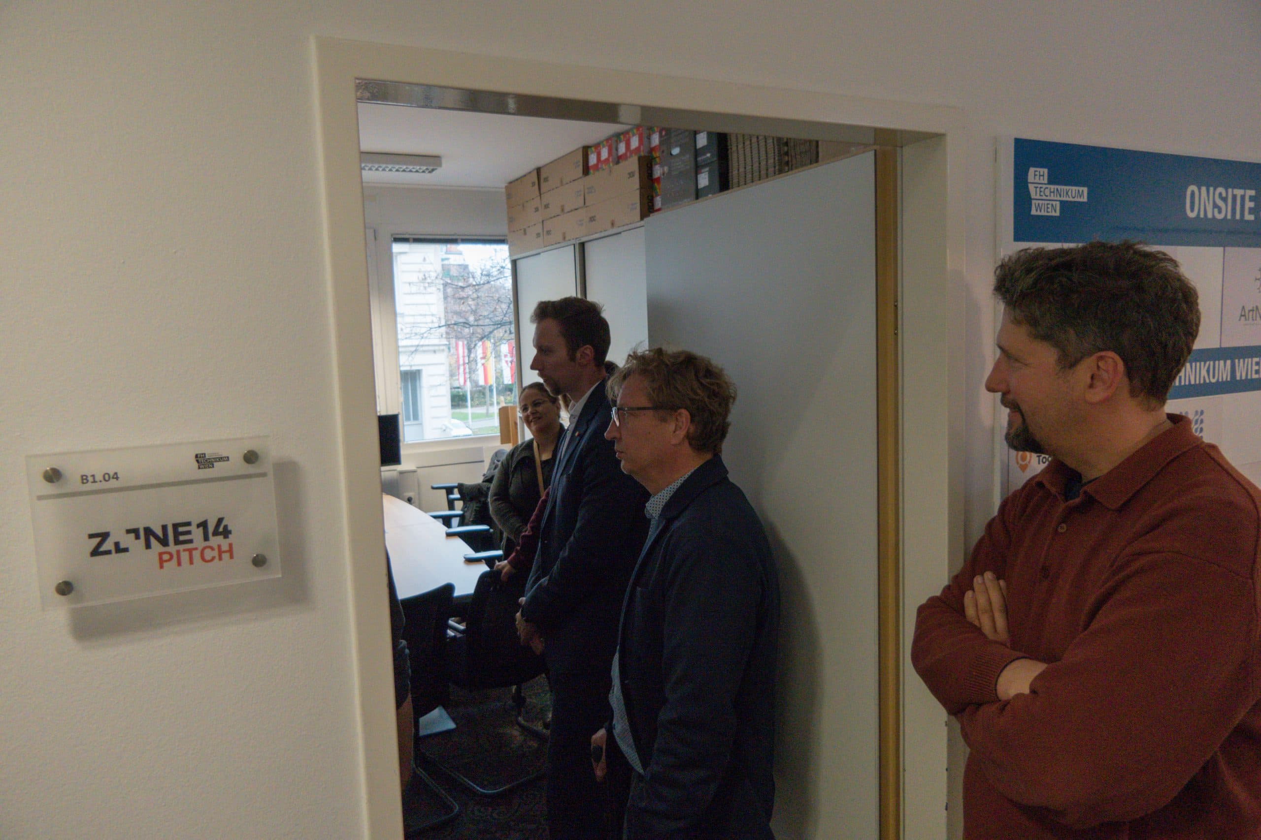 Vier Personen stehen in der Nähe der Tür eines Büros mit der Aufschrift 'ZONE14 PITCH' und blicken in einen Besprechungsraum mit Tischen, Stühlen und Regalen. Ein Fenster im Hintergrund zeigt Bäume und Gebäude im Freien.