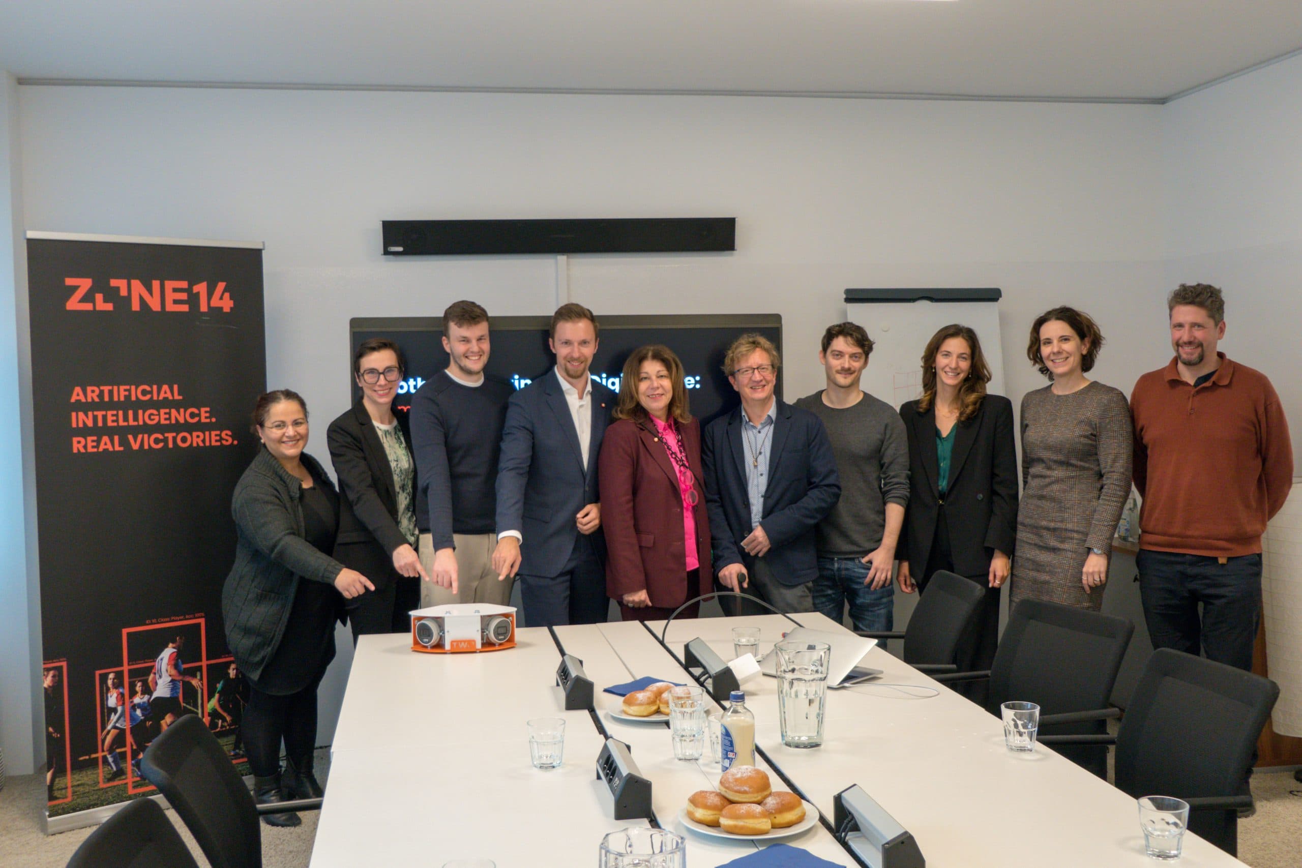 Neun Personen stehen lächelnd um einen Konferenztisch mit Gebäck und Getränken. Einige legen ihre Hände auf eine Schachtel. Auf einem Banner steht ZONE14 Artificial Intelligence. Echte Siege. im Hintergrund.