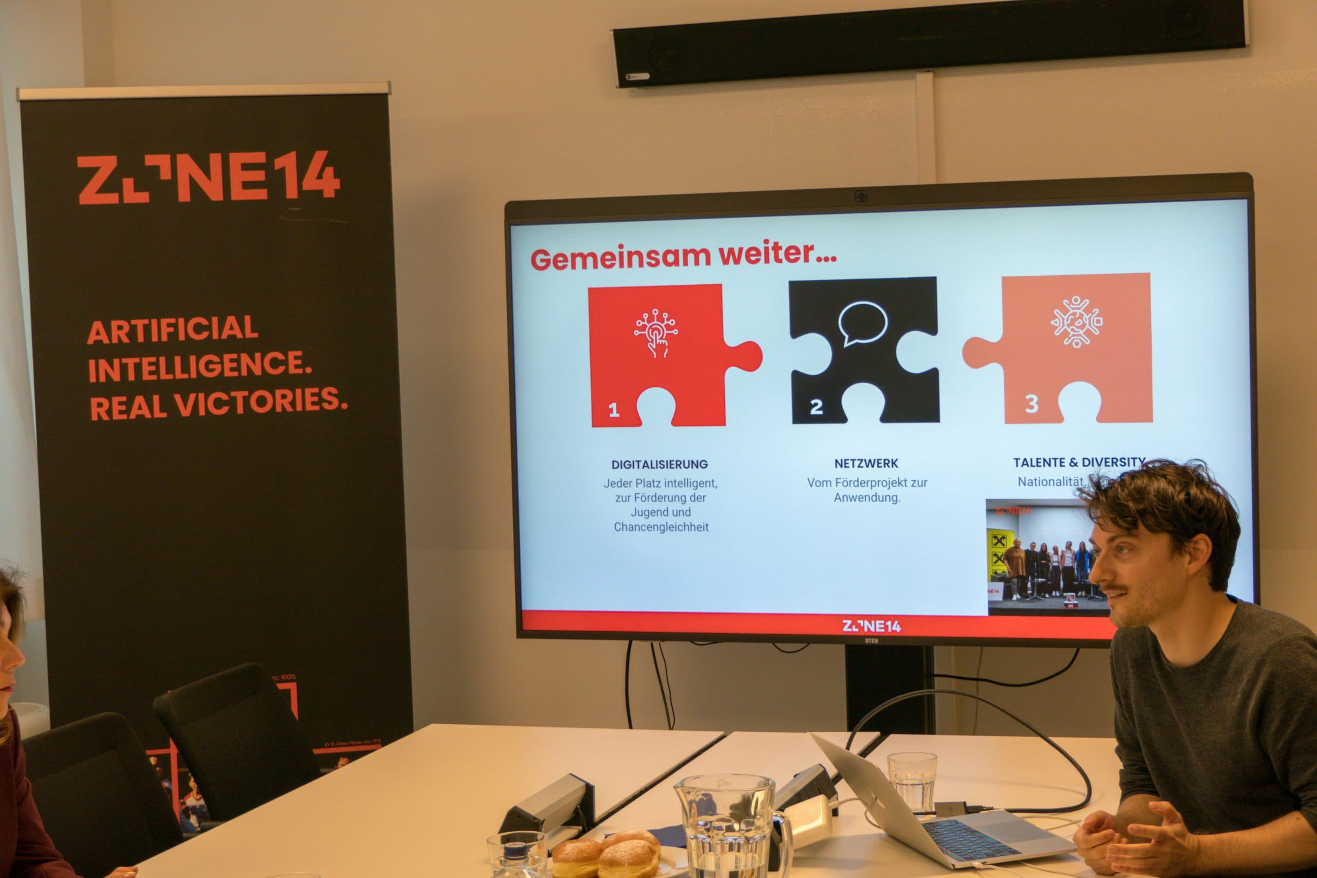 Ein Mann präsentiert eine Diashow in einem Konferenzraum. Auf dem Bildschirm erscheinen drei Puzzleteile mit den Bezeichnungen Digitalisierung, Netzwerk und Talente & Diversity. Auf der linken Seite ist ein Banner mit der Aufschrift Artificial Intelligence. Echte Siege.