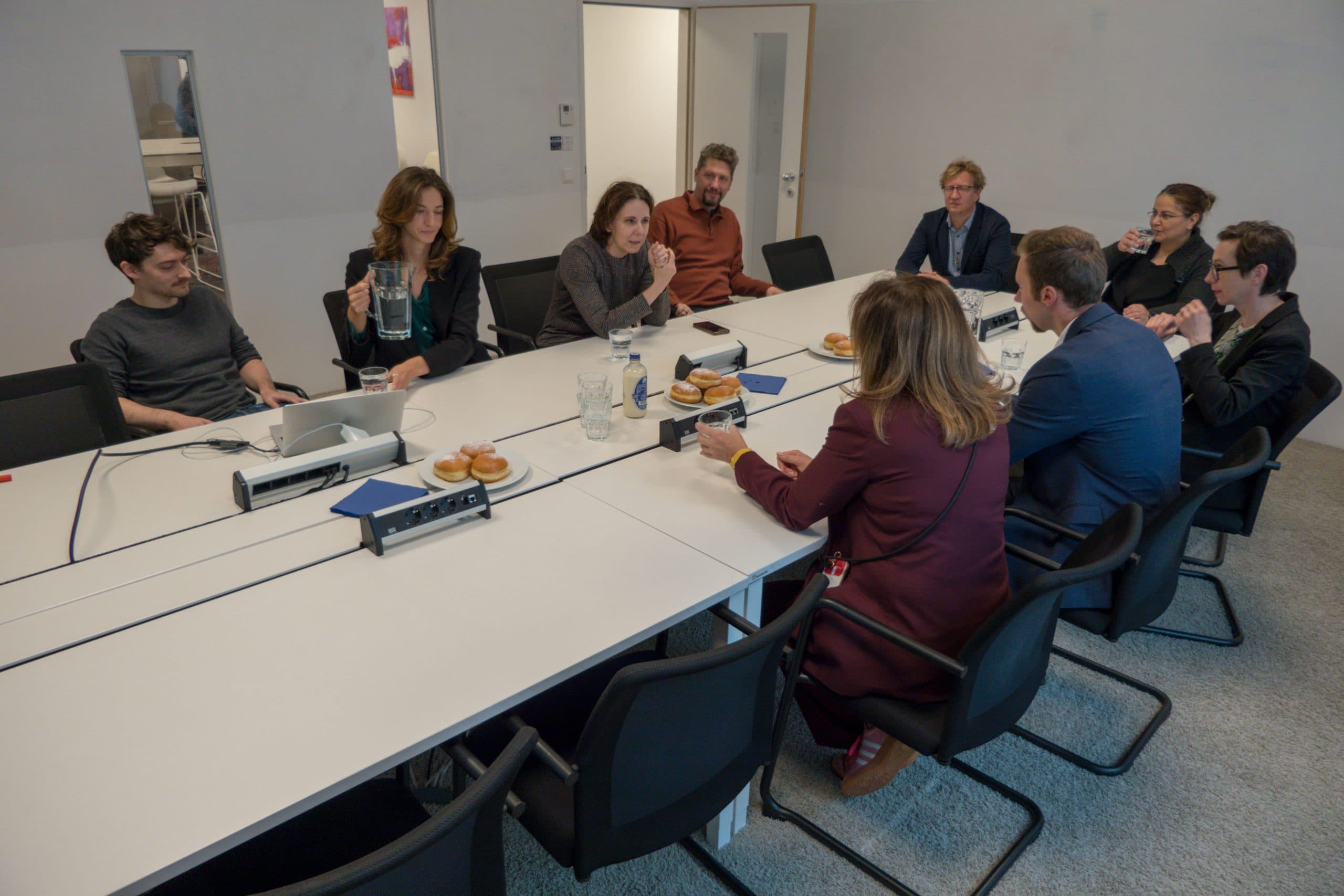 Eine Gruppe von neun Personen sitzt um einen großen Konferenztisch in einem Besprechungsraum und diskutiert. Auf dem Tisch stehen Laptops, Dokumente, Wasserkrüge und Teller mit Gebäck.