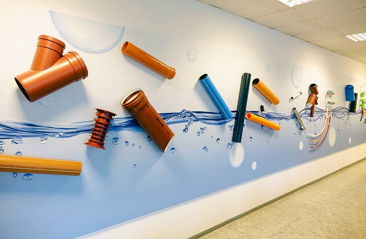 In einem Flur ist eine Wandmalerei mit blauem Wasser und Blasen zu sehen, auf der verschiedene bunte Kunststoffrohre und Armaturen in 3D montiert sind, was einen dynamischen, spielerischen Effekt erzeugt. Der Boden ist in hellen Farben gehalten und die Decke ist mit weißen Fliesen versehen.