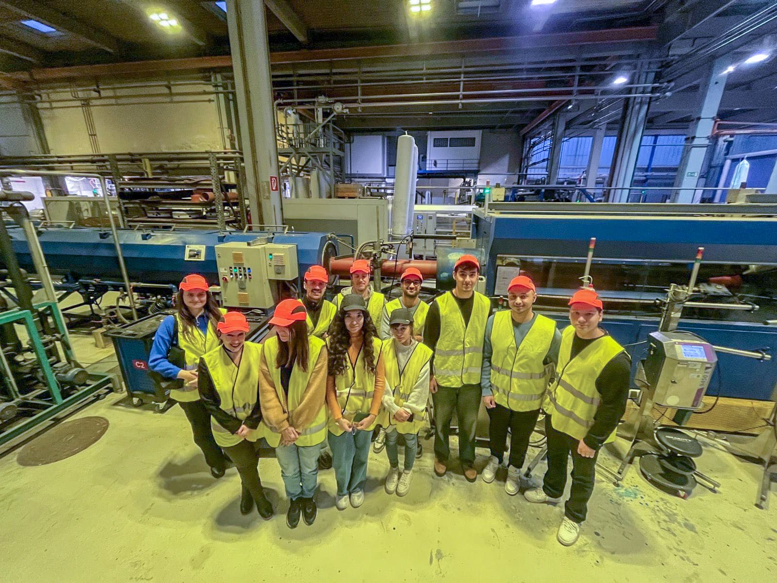 Eine Gruppe von Personen mit gelben Sicherheitswesten und roten Mützen steht in einer Industrieanlage zusammen und posiert für ein Foto. Sie sind umgeben von Maschinen und Rohren in einer gut beleuchteten, weitläufigen Fabrikanlage.