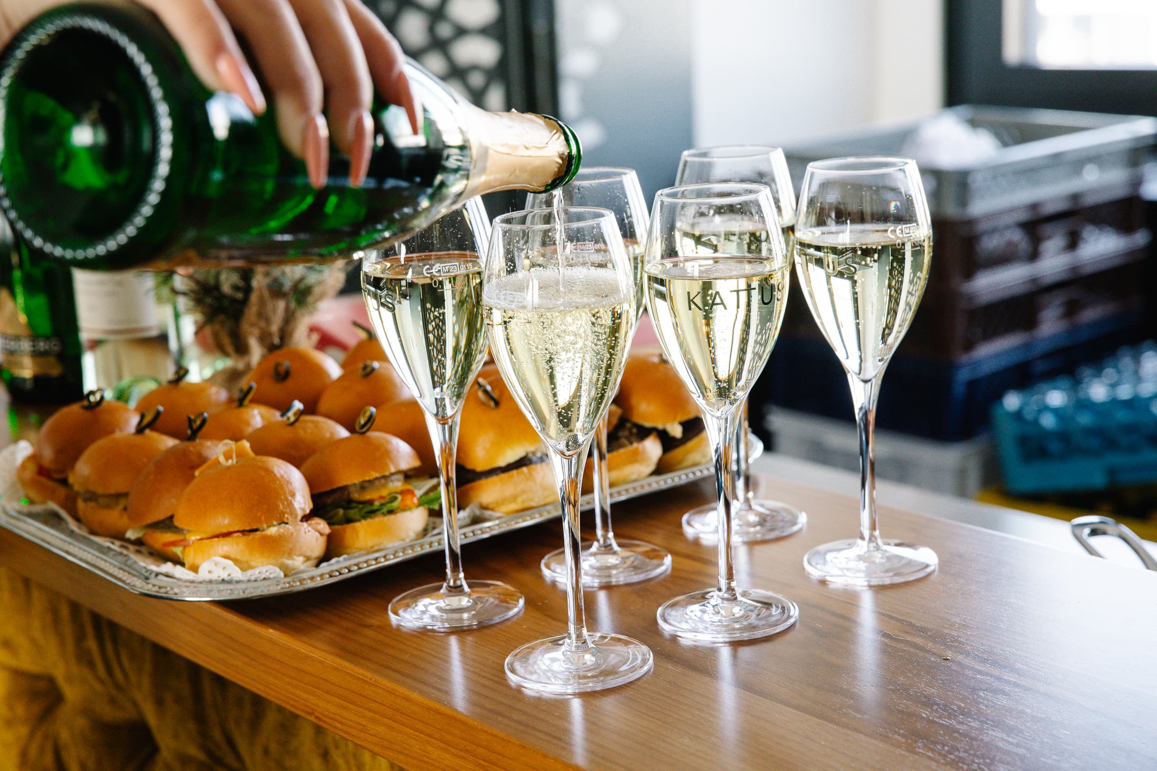 Eine Hand gießt Sekt in Gläser ein, die auf einem Tisch neben einem Tablett mit kleinen Sandwiches stehen. Die Umgebung deutet auf ein feierliches oder gesellschaftliches Ereignis hin.