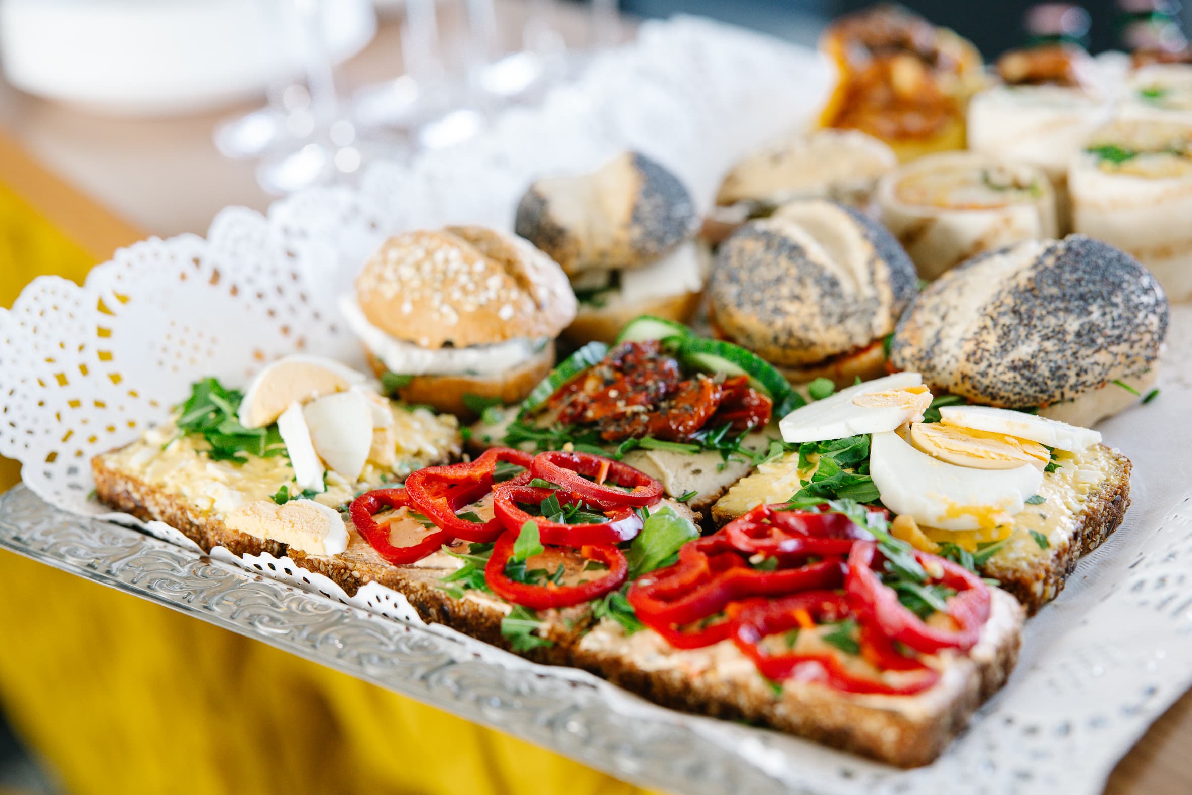 Ein Tablett mit verschiedenen belegten Brötchen, belegt mit in Scheiben geschnittenen Eiern, rotem Paprika, Grünzeug und Mohnbrötchen, präsentiert auf einem dekorativen Deckchen aus Papier.