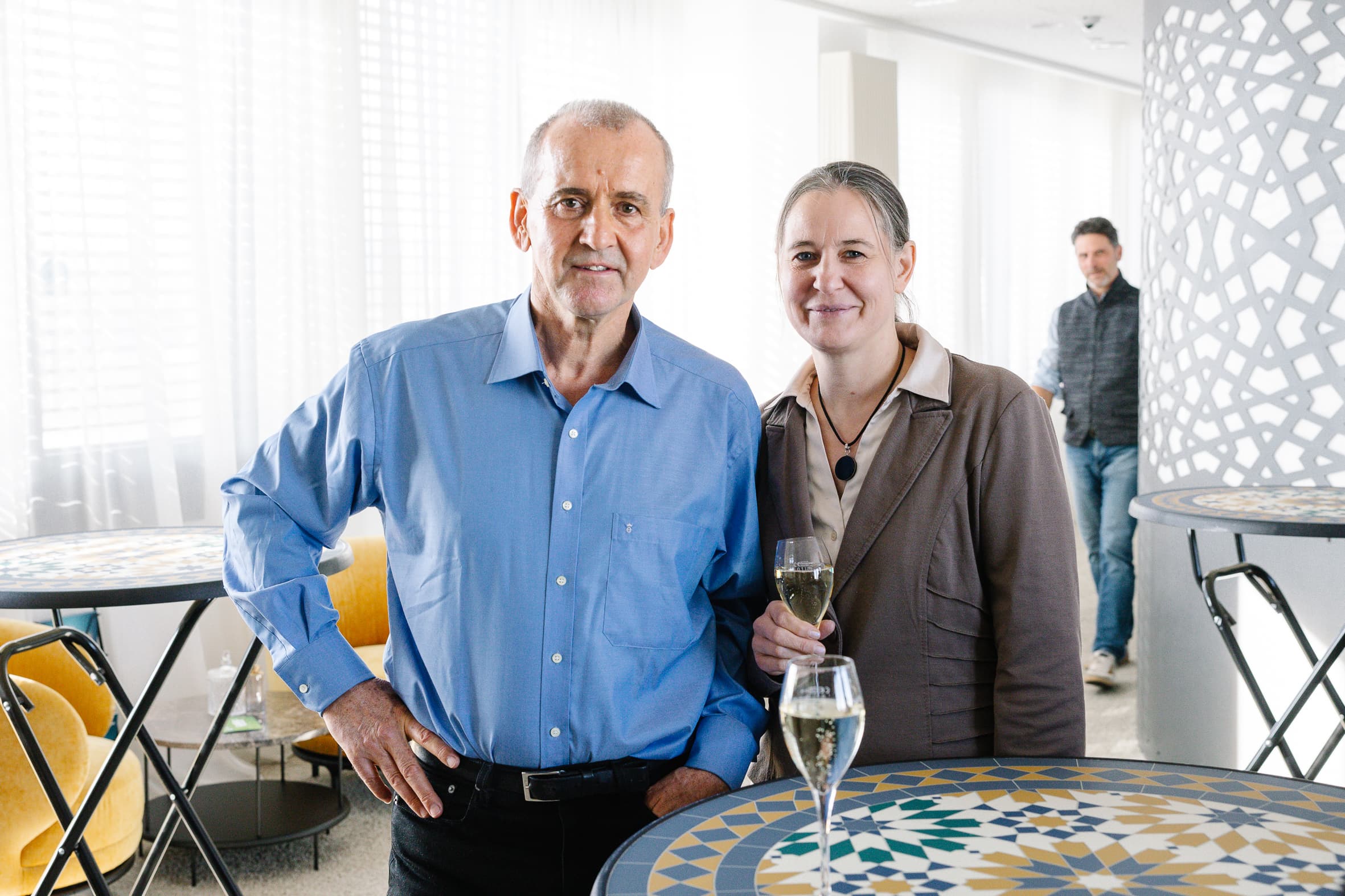 Ein Mann in einem blauen Hemd und eine Frau in einer braunen Jacke stehen an einem gemusterten Tisch, lächeln in die Kamera und halten Getränke in der Hand. Eine weitere Person ist im Hintergrund in der Nähe einer modernen, geometrischen Wand zu sehen.