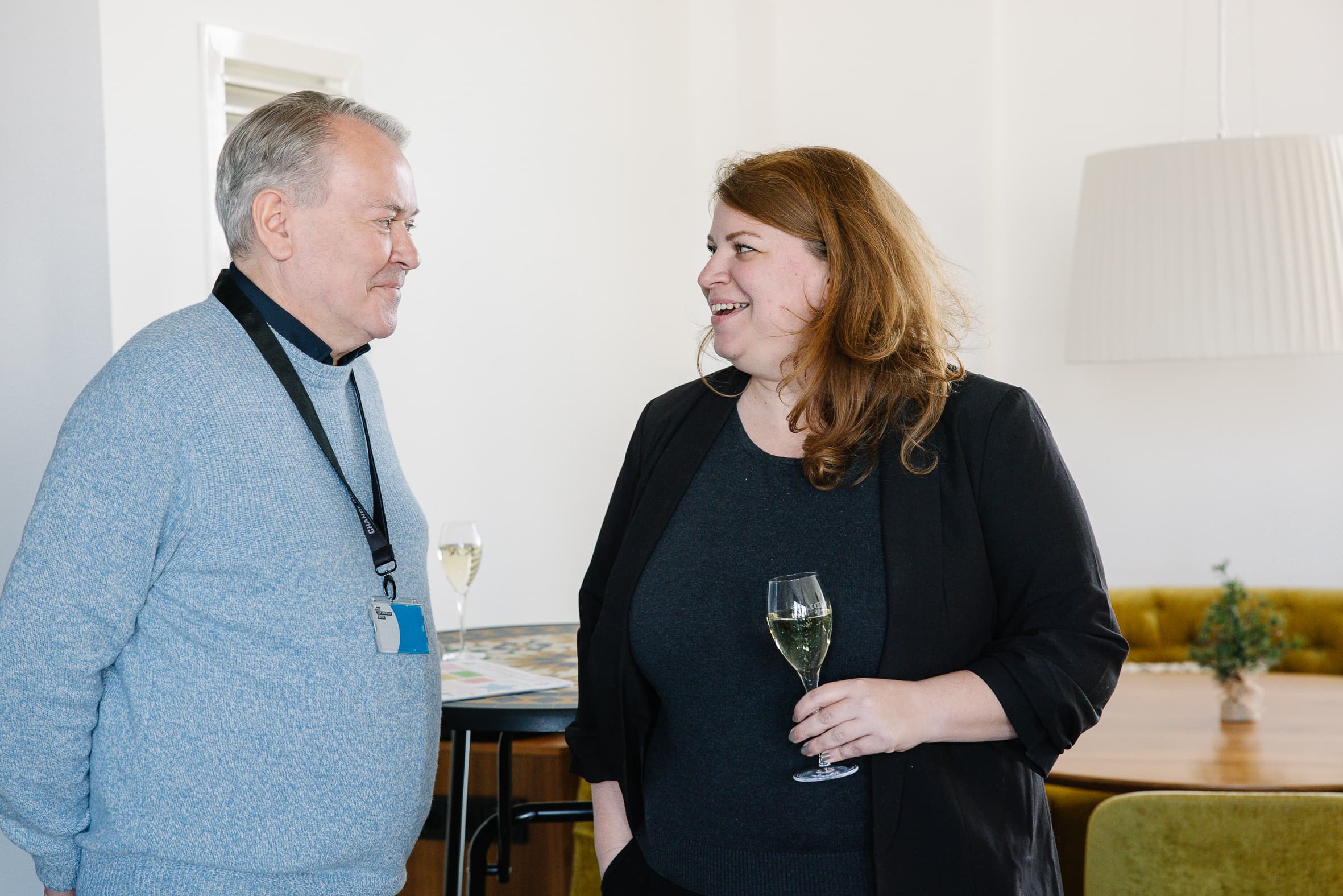 Ein Mann und eine Frau stehen in einem Haus, lächeln und unterhalten sich. Die Frau hält ein Glas Weißwein in der Hand. Es scheint, als befänden sie sich bei einem gesellschaftlichen oder beruflichen Treffen in einem gut beleuchteten, modern eingerichteten Raum.