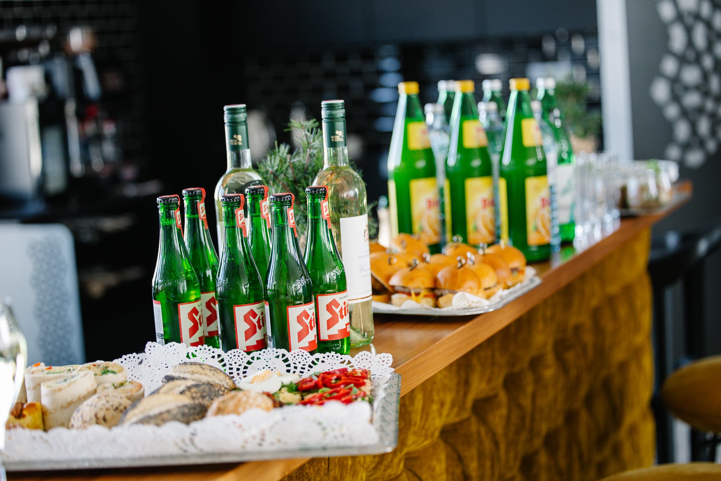 Auf einer Holztheke stehen grüne Glasflaschen, eine Weinflasche, belegte Brötchen, Mini-Burger, Gläser und Teller mit Vorspeisen, die für ein Buffet oder einen Empfang hübsch angeordnet sind.