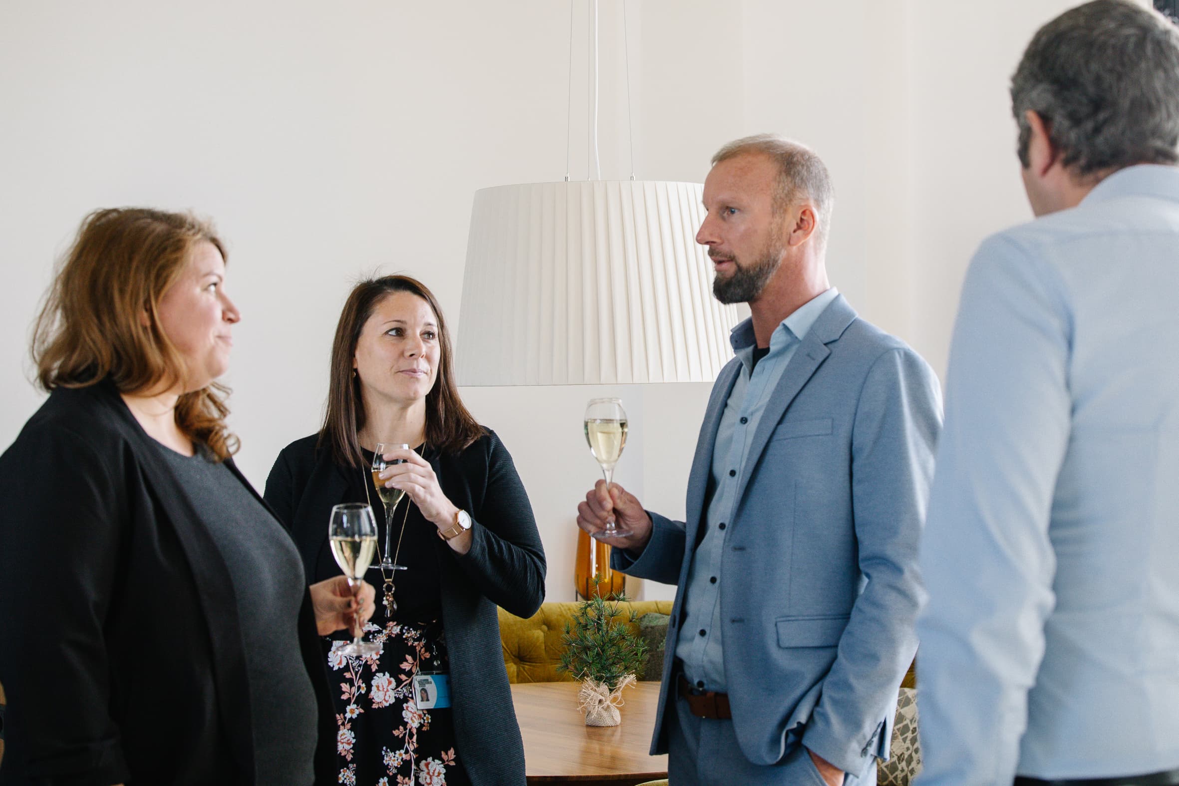 Vier Personen in Geschäftskleidung stehen in einem Innenraum zusammen, halten Gläser mit Champagner in der Hand und unterhalten sich. Eine große weiße Lampe hängt im Hintergrund, und eine kleine Pflanze steht auf einem Tisch in der Nähe.