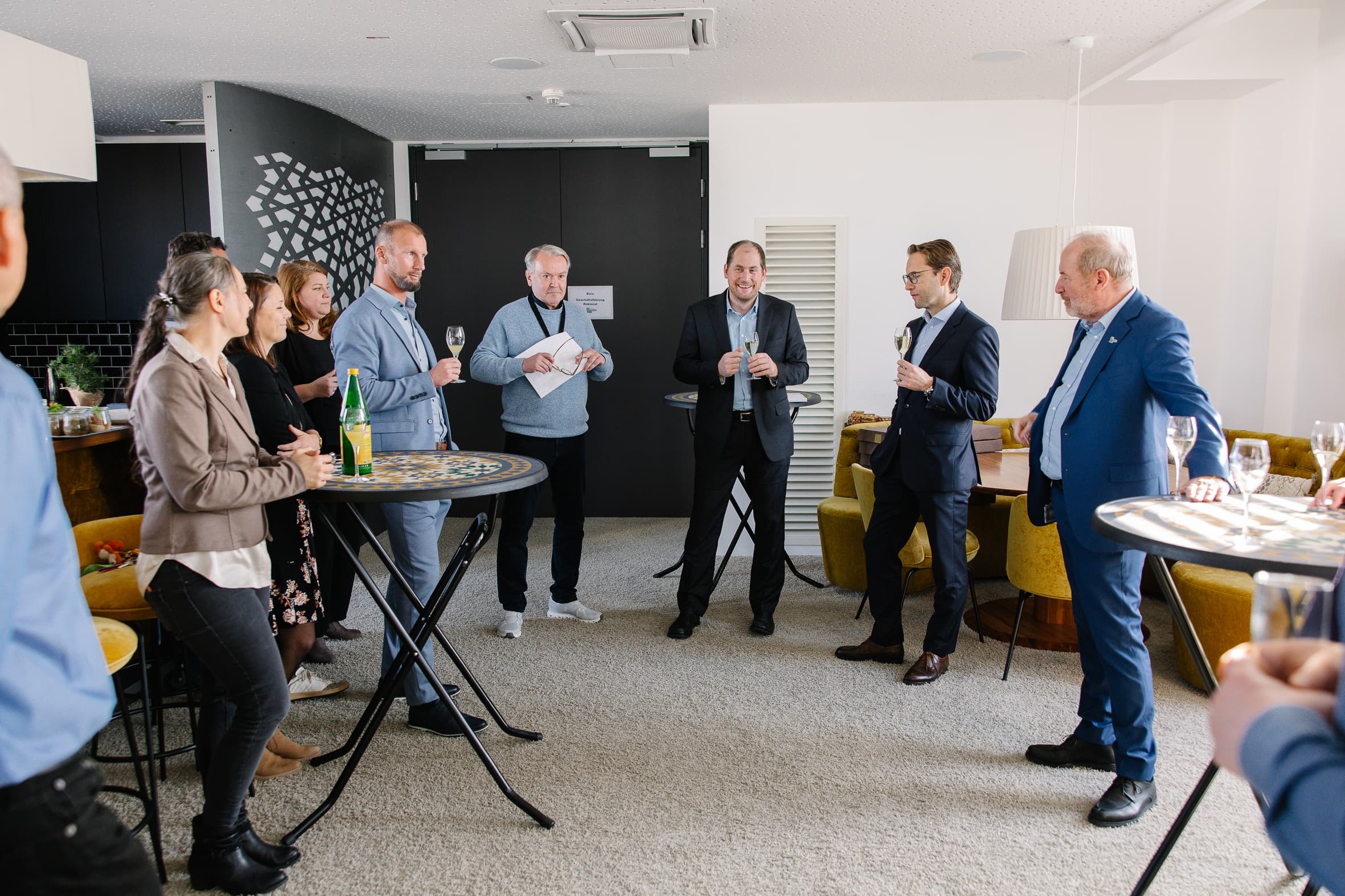 Eine Gruppe von Menschen in Geschäftskleidung steht in einer modernen Lounge, unterhält sich und hält Getränke an Stehtischen. Einige lächeln und hören einem Mann zu, der eine Zeitung hält. Die Atmosphäre wirkt locker und gesellig.