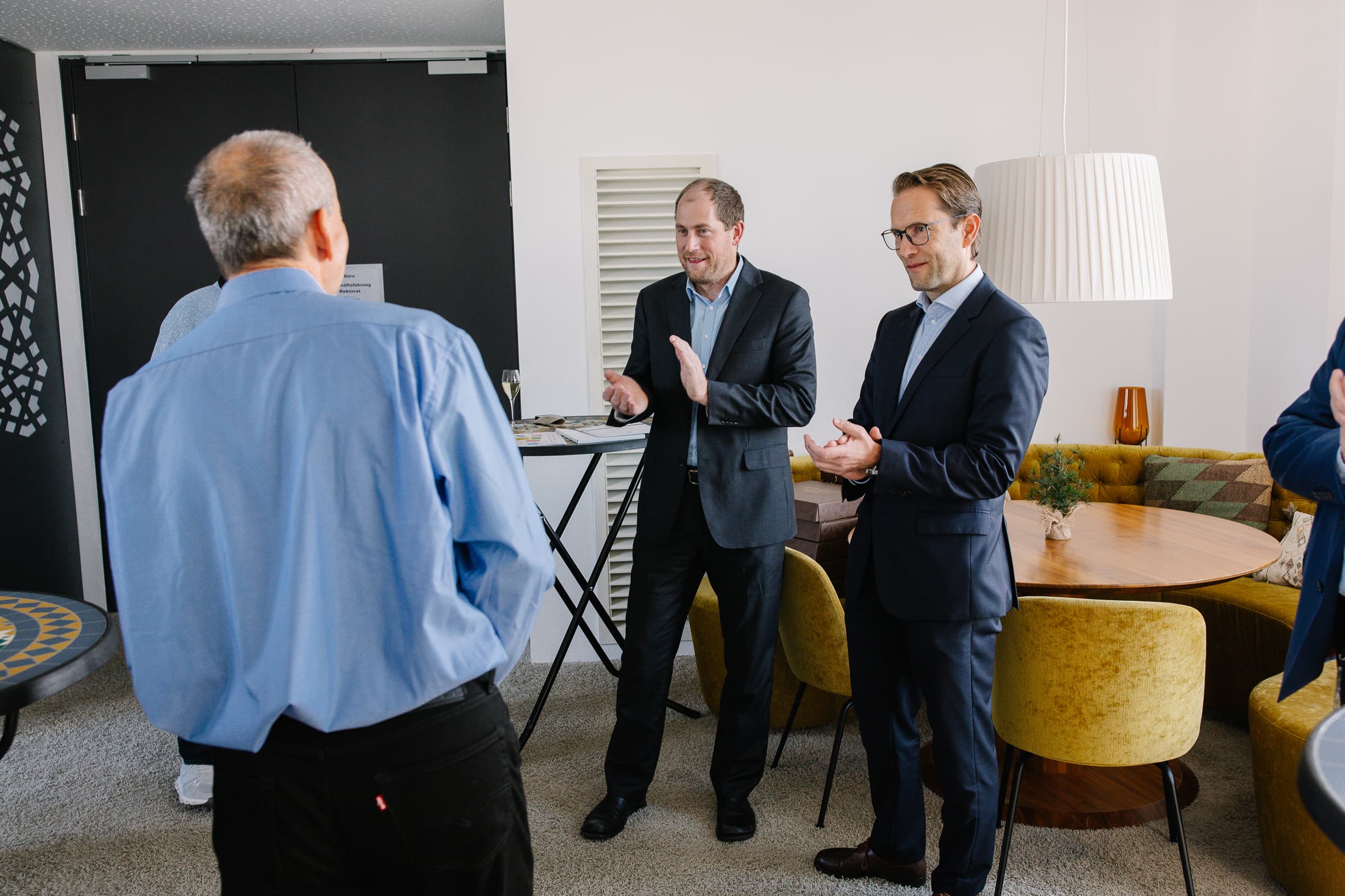 Drei Männer in Geschäftskleidung stehen in einer modernen Bürolounge und unterhalten sich, wobei ein Mann von der Kamera wegschaut und die beiden anderen lächeln und gestikulieren, während sie sprechen. Im Hintergrund stehen ein runder Tisch und goldene Stühle.