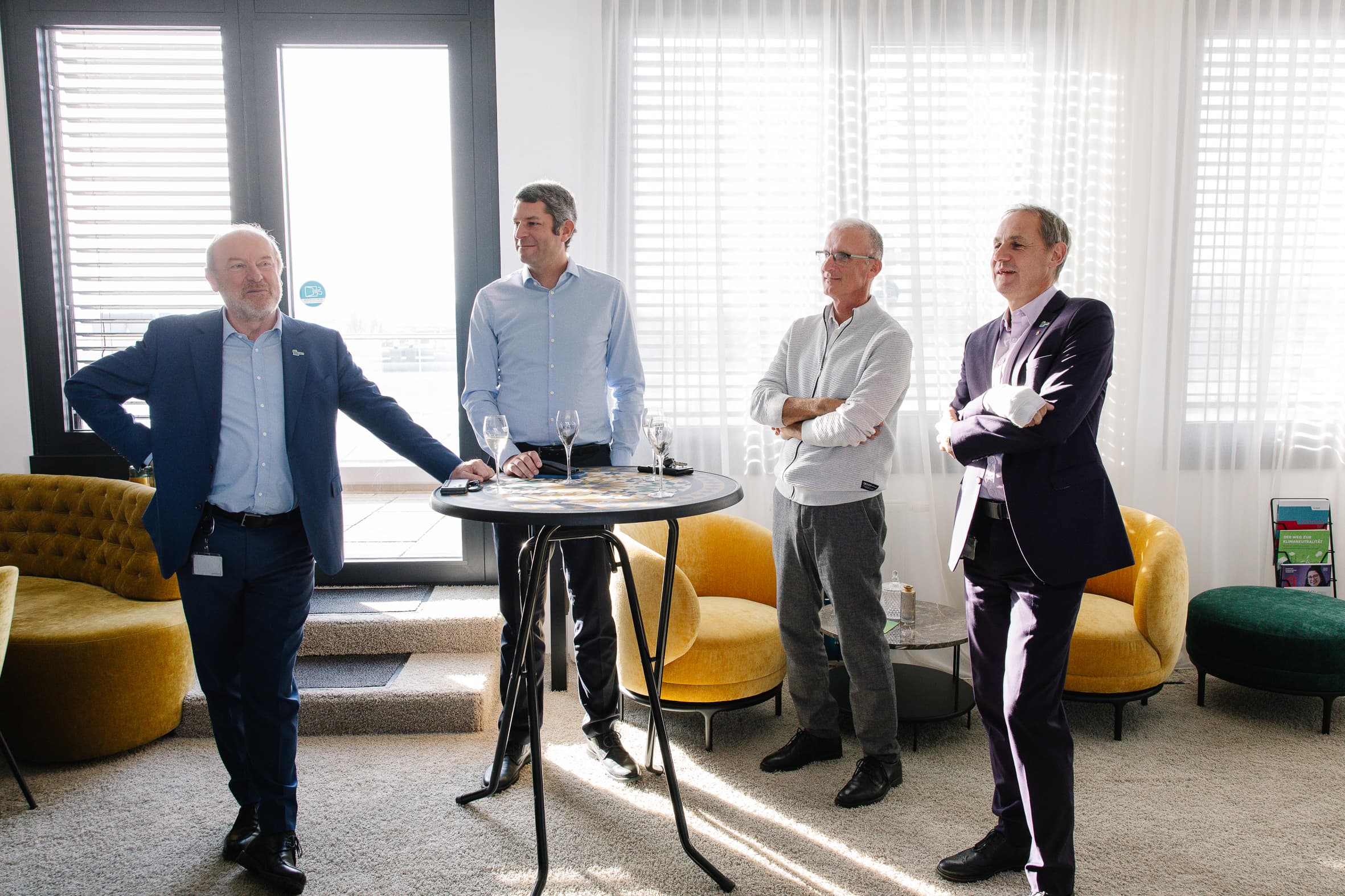 Vier Männer in Geschäftskleidung stehen um einen hohen runden Tisch mit Getränken in einem hellen, modernen Raum. Sonnenlicht strömt durch große Fenster mit Jalousien. Im Hintergrund stehen zwei senfgelbe Stühle und einige Zeitschriften.