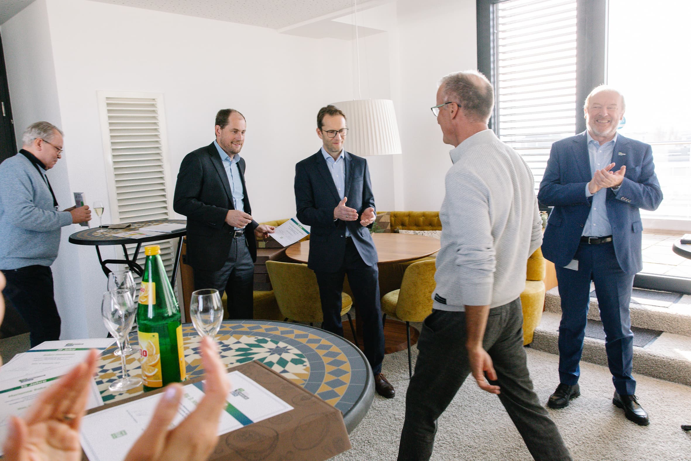 Fünf Männer in Geschäftskleidung versammeln sich in einem hellen Raum, einige klatschen und lächeln. Ein Mann geht auf die Gruppe zu, während auf den Tischen in der Nähe Wasserflaschen, Gläser und Dokumente stehen. Die Atmosphäre wirkt freundlich und feierlich.