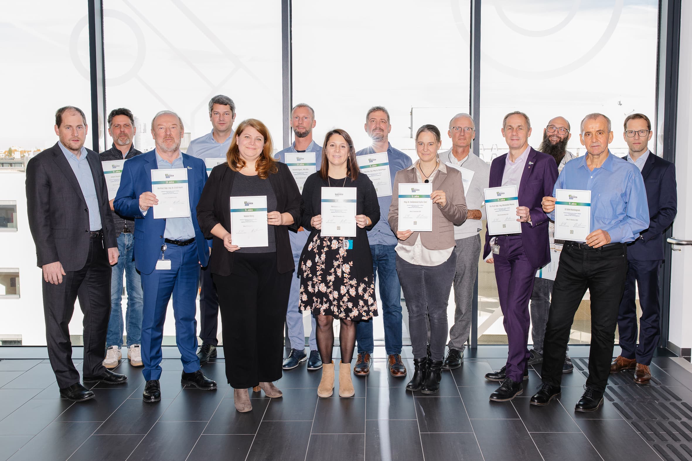 Eine Gruppe von fünfzehn Personen steht in einem Innenraum vor großen Fenstern, hält Urkunden in der Hand und lächelt in die Kamera. Sie sind in Business-Kleidung gekleidet.