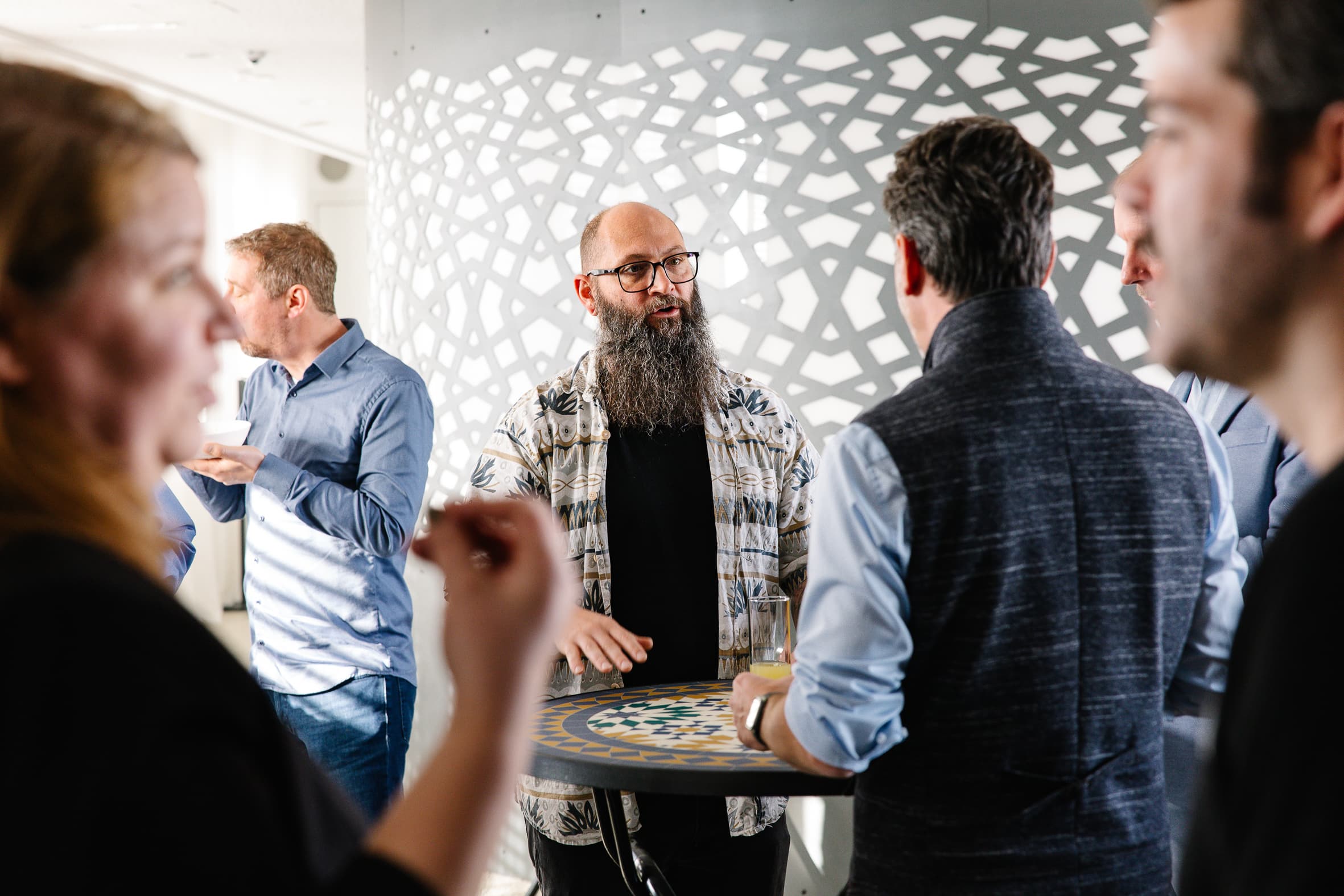 Eine Gruppe von Menschen steht um einen hohen Tisch herum und unterhält sich bei einem geselligen Beisammensein. Ein Mann mit langem Bart und Brille steht im Mittelpunkt, während andere in einem hell erleuchteten Raum plaudern und gestikulieren.