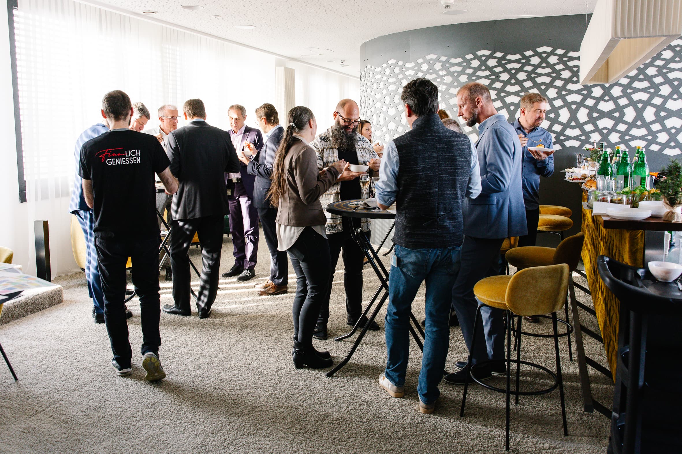Eine Gruppe von Menschen versammelt sich um Stehtische in einem hellen Raum, auf dem Tresen stehen Getränke und Speisen, was auf ein zwangloses Networking oder eine gesellschaftliche Veranstaltung hindeutet. Durch große Fenster strömt Sonnenlicht.