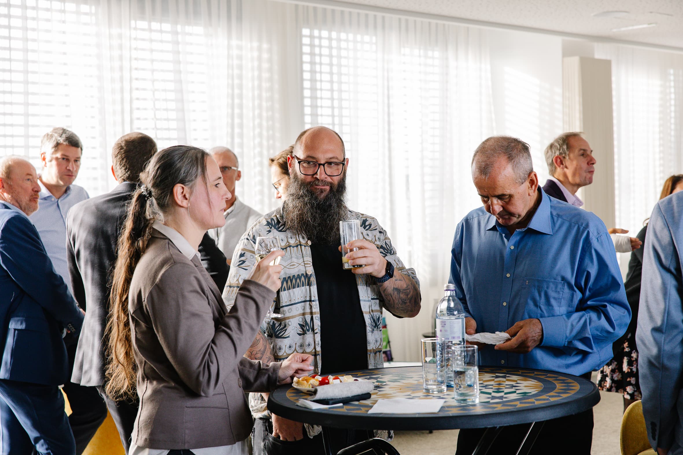 Eine Gruppe von Menschen steht bei einem gesellschaftlichen Ereignis um einen runden Tisch herum und unterhält sich; eine Frau hält einen Teller und eine Gabel, ein bärtiger Mann mit Brille hält ein Getränk, und ein älterer Mann liest seinen Teller. Andere unterhalten sich im Hintergrund.