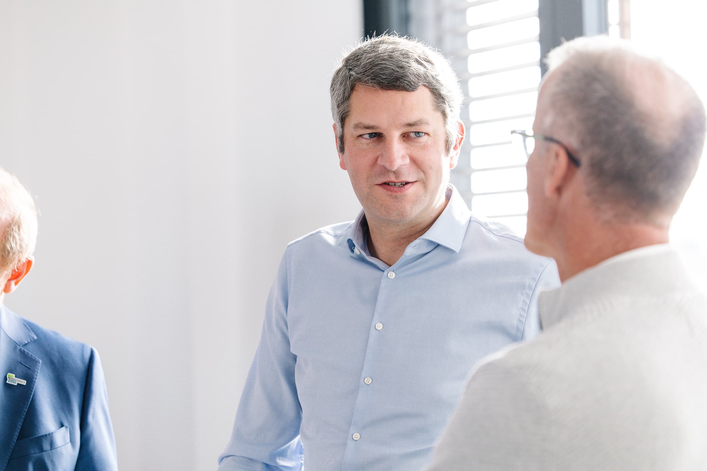 Ein Mann in einem hellblauen Hemd unterhält sich mit einem anderen Mann mit Brille, der einen hellen Pullover trägt. Sie befinden sich in einem Innenraum in der Nähe eines Fensters mit Jalousien, durch das Sonnenlicht eindringt. Eine dritte Person ist teilweise sichtbar.