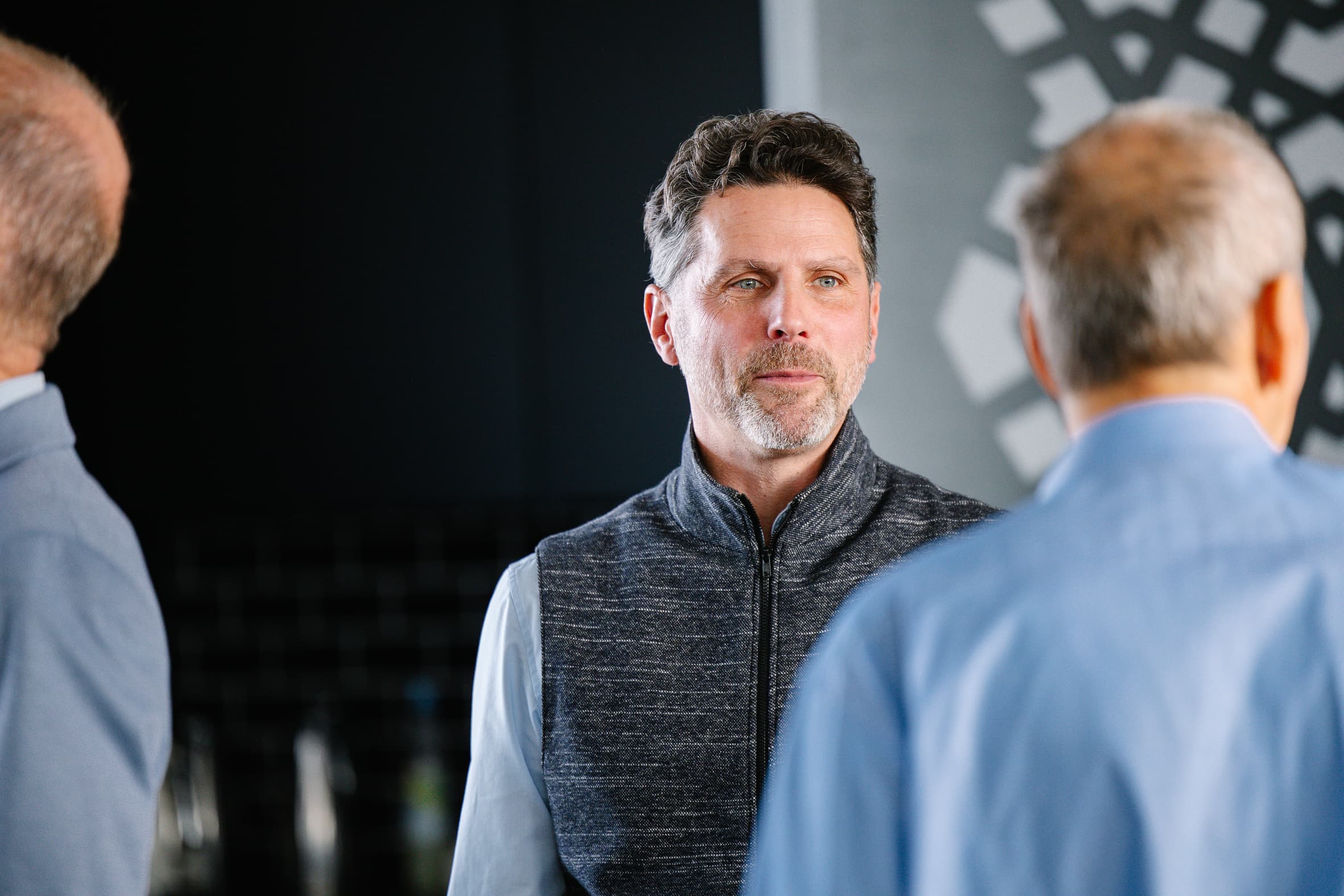 Ein Mann mit kurzen braunen Haaren und einer grauen Weste unterhält sich mit zwei anderen Männern, die alle blaue Hemden tragen, in einem Innenraum mit dunklem Hintergrund.