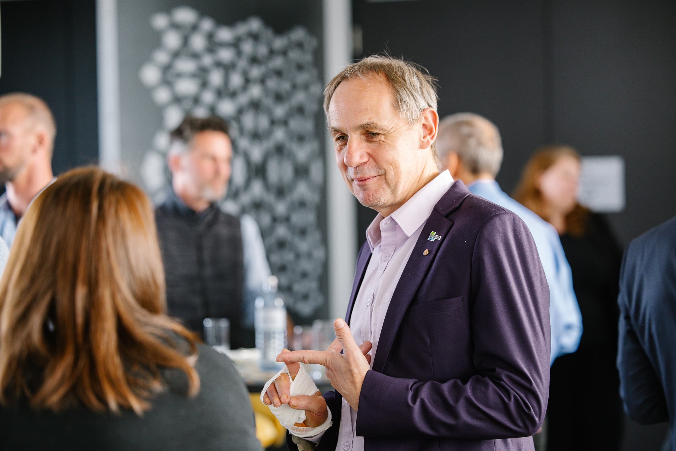 Ein Mann in einem violetten Blazer gestikuliert, während er mit einer Frau bei einem geselligen Beisammensein spricht. Mehrere Personen unterhalten sich im unscharfen Hintergrund. Die Umgebung scheint ein Innenraum zu sein.
