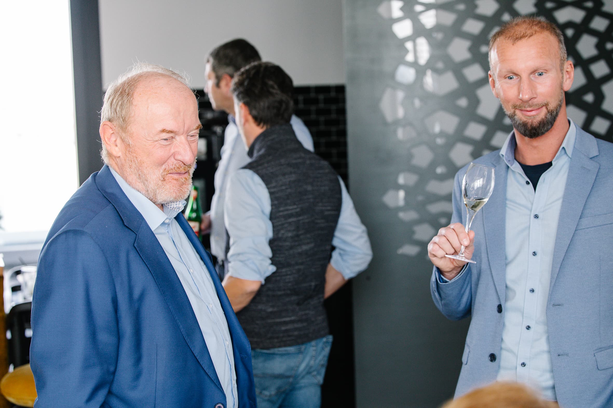 Zwei Männer in blauen Anzügen stehen in einem Innenraum bei einer gesellschaftlichen Veranstaltung. Ein Mann hält ein leeres Weinglas und lächelt, während der andere in die Kamera schaut. Im Hintergrund sind Menschen zu sehen, und die Atmosphäre ist locker.