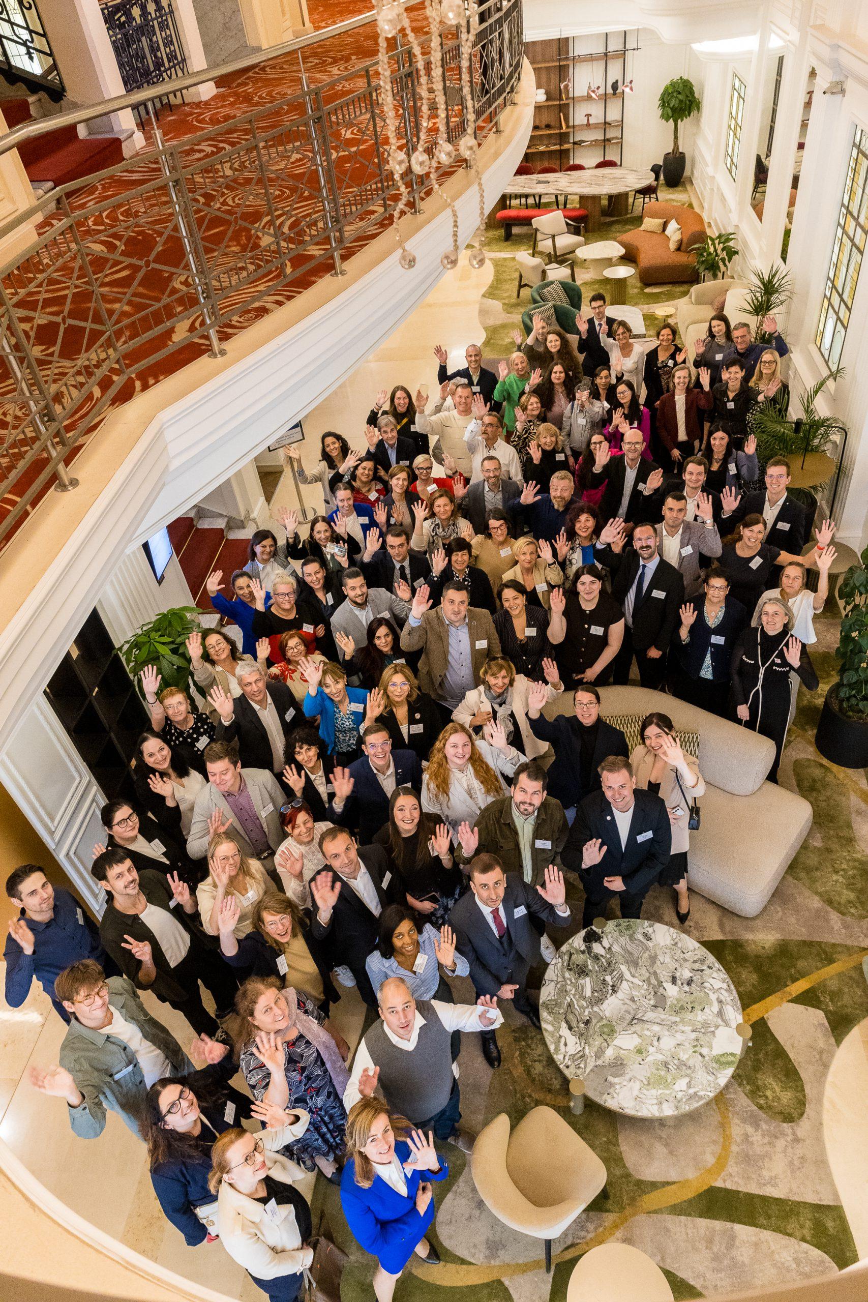 Eine große Gruppe von Menschen in Geschäftskleidung steht in einer Hotellobby zusammen, schaut auf und lächelt in die Kamera, viele mit einer zum Gruß erhobenen Hand, umgeben von modernen Möbeln und dekorativen Pflanzen.