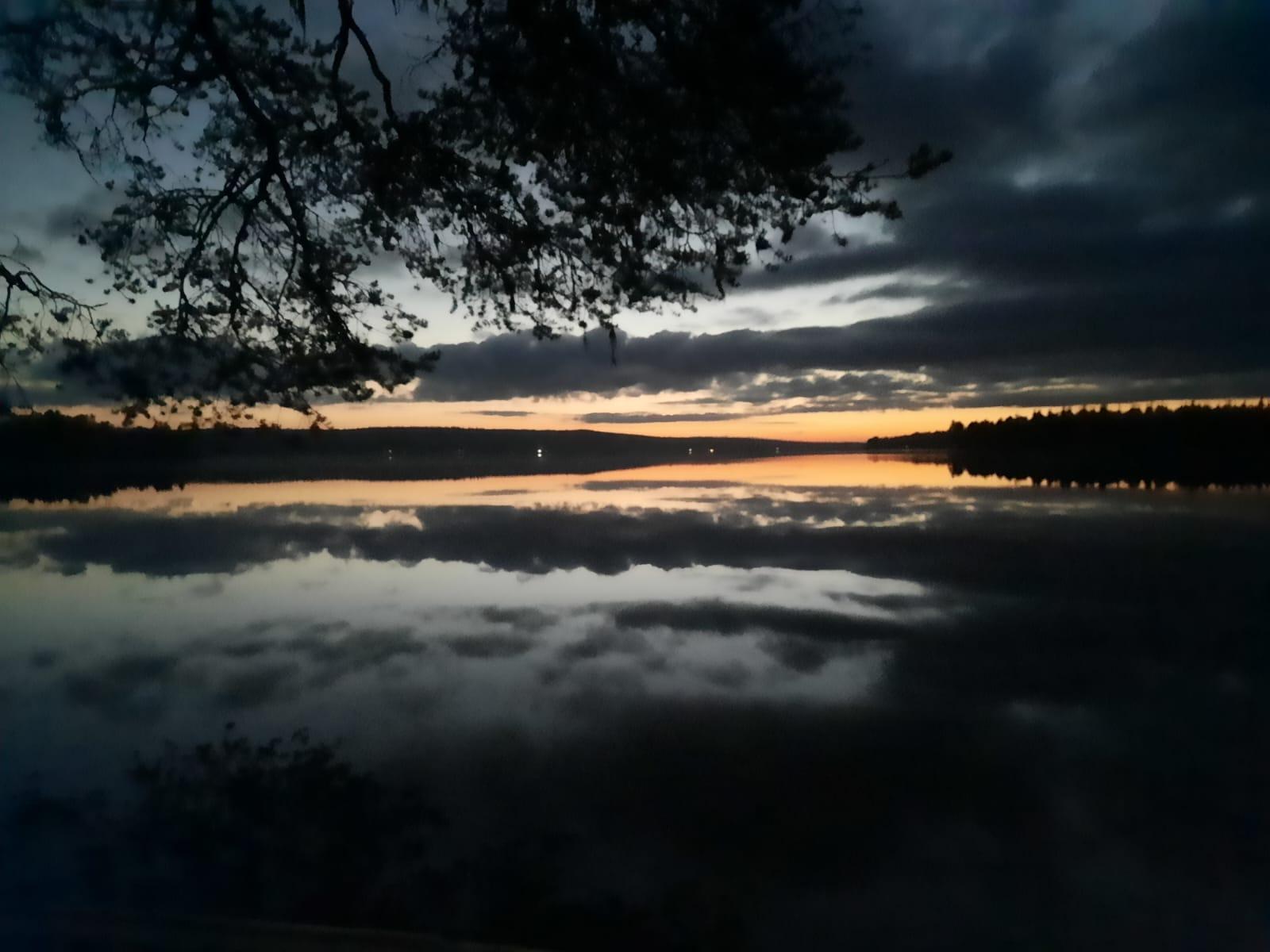 Ein ruhiger See bei Sonnenuntergang, mit dunklen Wolken und Bäumen, die sich gegen den orange-blauen Himmel abheben, und einer Landschaft, die sich auf dem stillen Wasser spiegelt. Zweige rahmen den oberen Teil des Bildes ein.