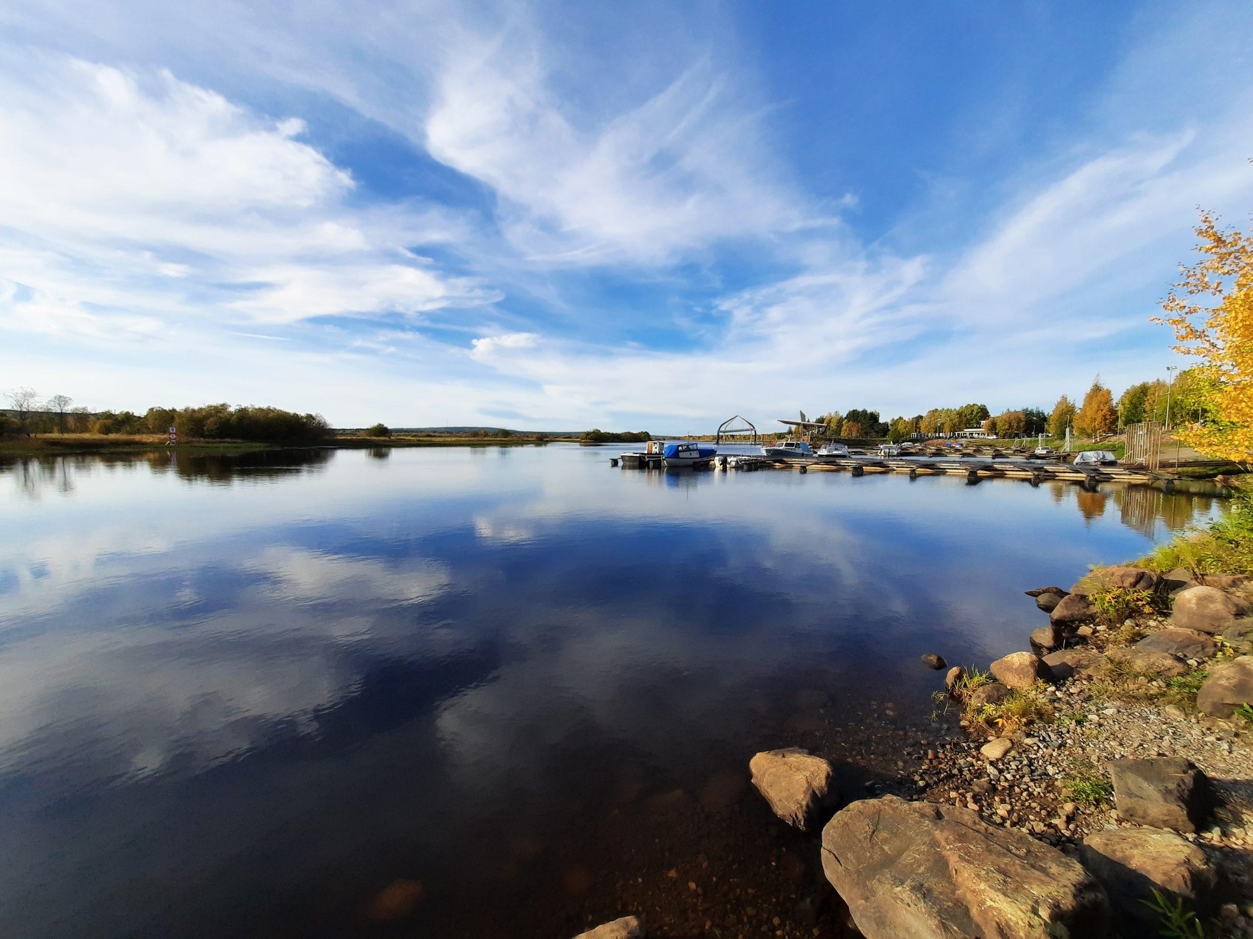 Ein ruhiger See mit einem felsigen Ufer, einem Steg mit Booten und Bäumen mit Herbstlaub unter einem blauen Himmel mit Wolkenfetzen, die sich im Wasser spiegeln.