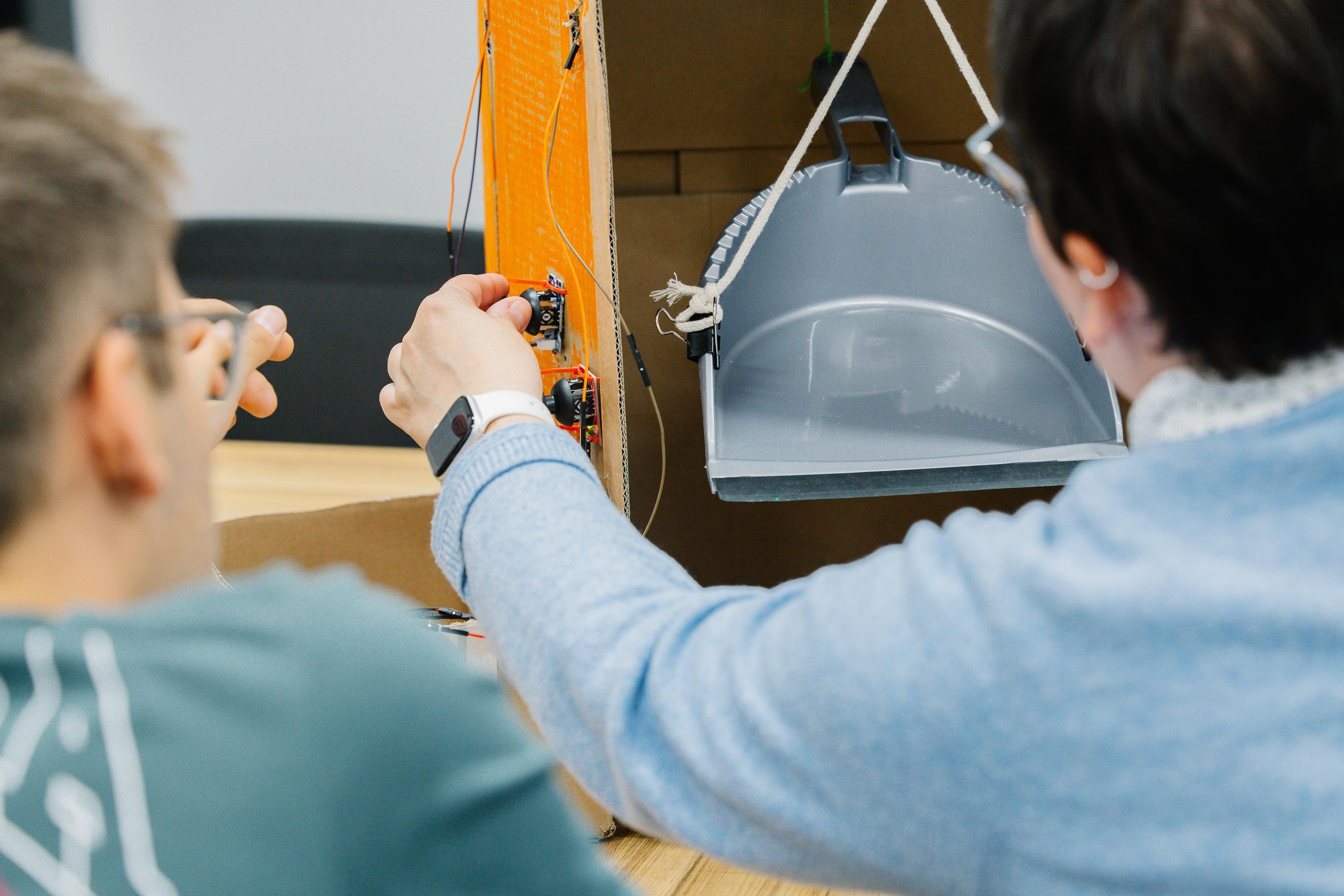 Zwei Personen arbeiten gemeinsam an einem technischen Projekt, bei dem sie elektronische Komponenten einstellen, die an einer Pappstruktur befestigt sind, in der eine graue Schaufel oder ein Behälter hängt.
