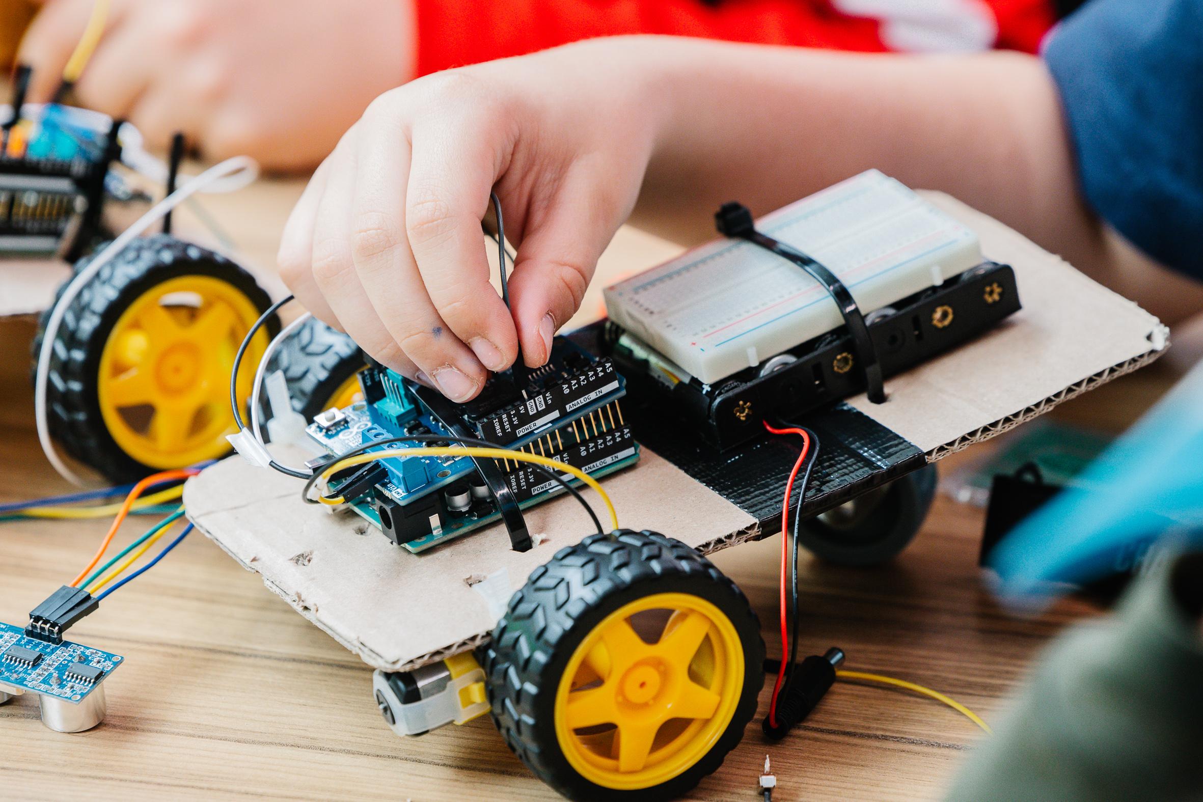 Nahaufnahme einer Kinderhand, die elektronische Komponenten an einem kleinen selbstgebauten Roboterauto mit gelben Rädern, Drähten, einer Lochrasterplatine und einem Mikrocontroller auf einem Holztisch zusammenbaut.