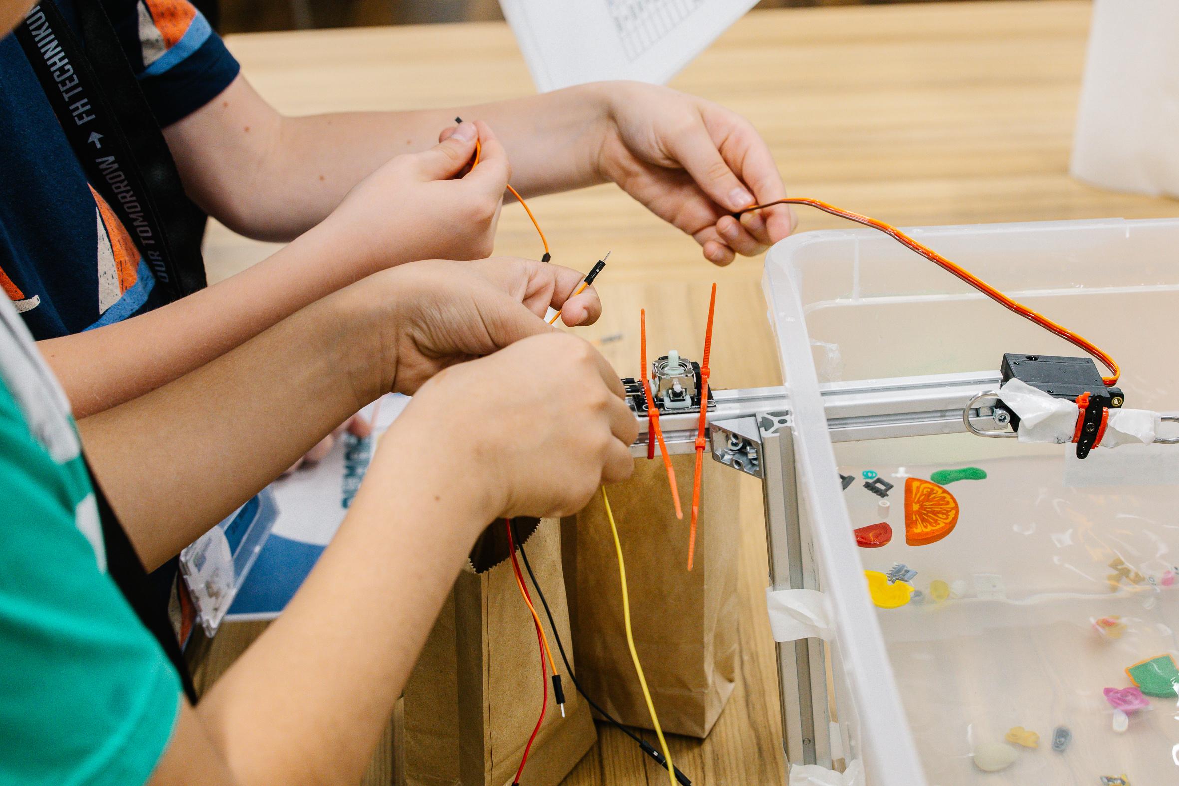 Drei Kinderhände arbeiten zusammen, um Drähte mit einem kleinen Roboter zu verbinden, der neben einem durchsichtigen Behälter mit bunten Gegenständen auf einem Holztisch steht.