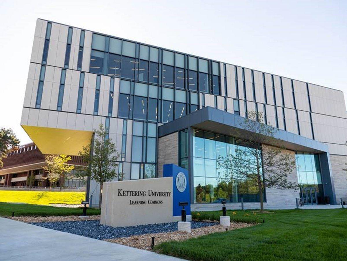 Ein modernes Gebäude aus Glas und Metall mit großen Fenstern an der Kettering University, das als Learning Commons bezeichnet wird, umgeben von grünem Gras und ein paar jungen Bäumen.