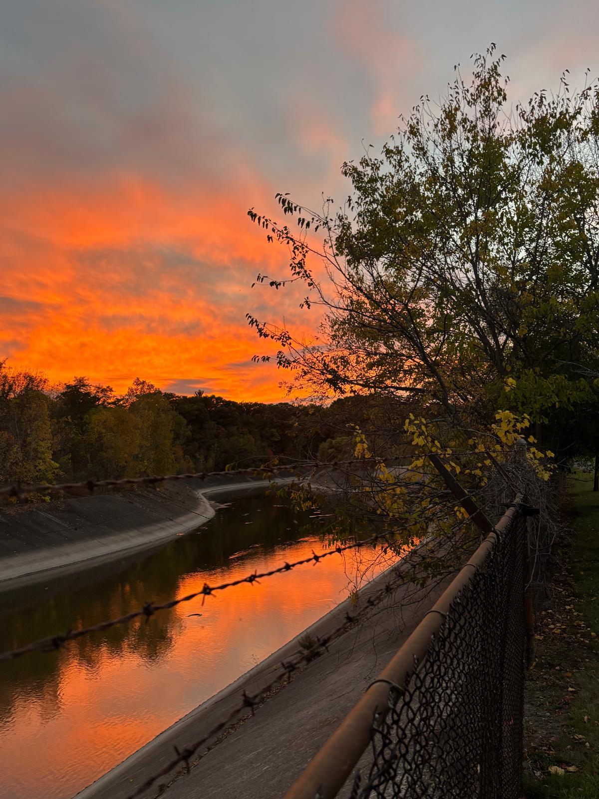 Ein leuchtend oranger und rosafarbener Sonnenuntergang spiegelt sich auf dem ruhigen Wasser eines von Bäumen gesäumten Kanals. Auf der rechten Seite verläuft ein Maschendrahtzaun, und der Himmel ist mit dramatischen Wolken gefüllt.