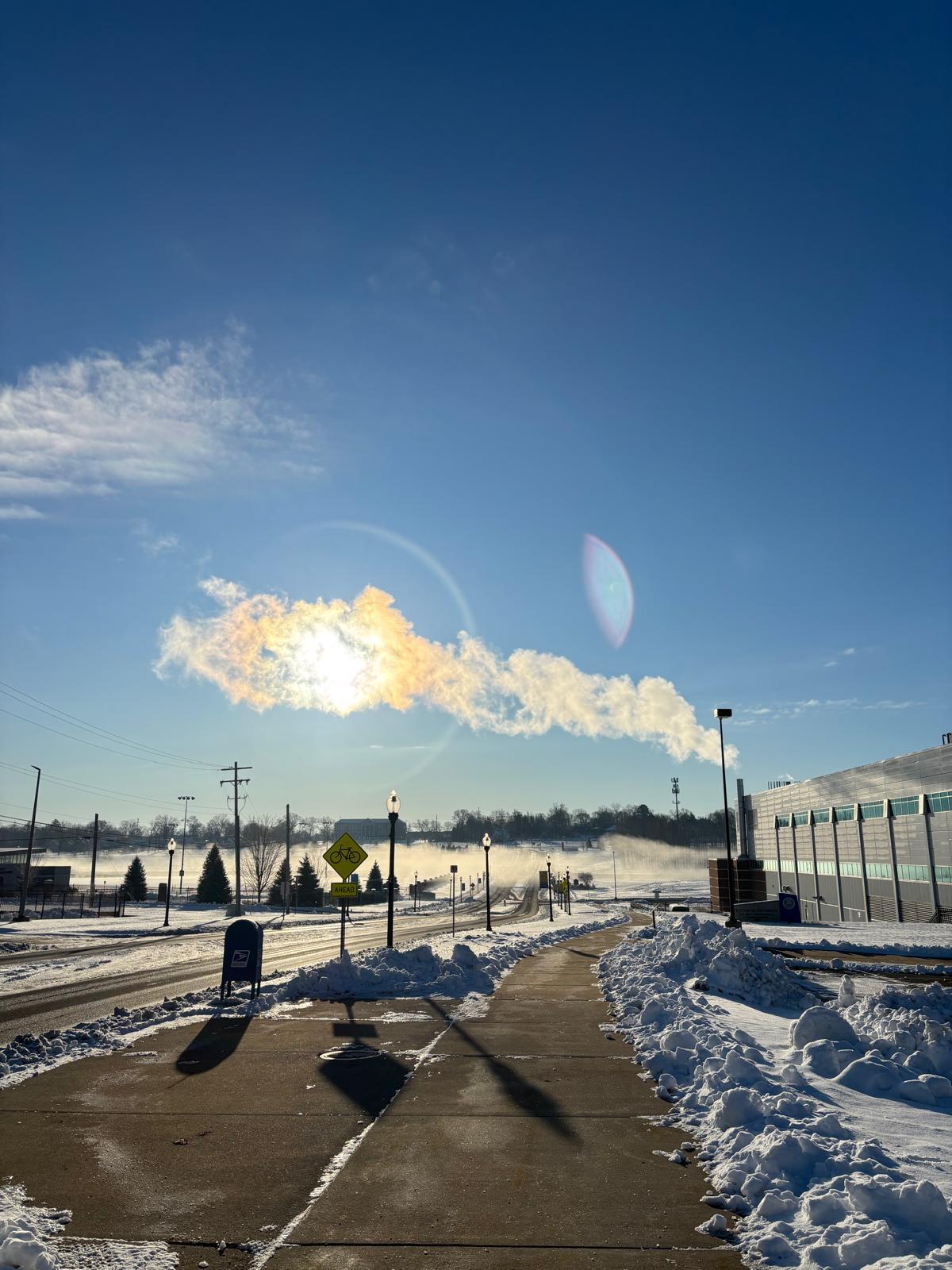 Ein sonniger Wintertag, an dem der Boden und die Gehwege mit Schnee bedeckt sind, klarer blauer Himmel, Dampf oder Rauch steigt in der Ferne auf, und ein Briefkasten und Straßenschilder sind entlang einer Straße neben einem modernen Gebäude zu sehen.
