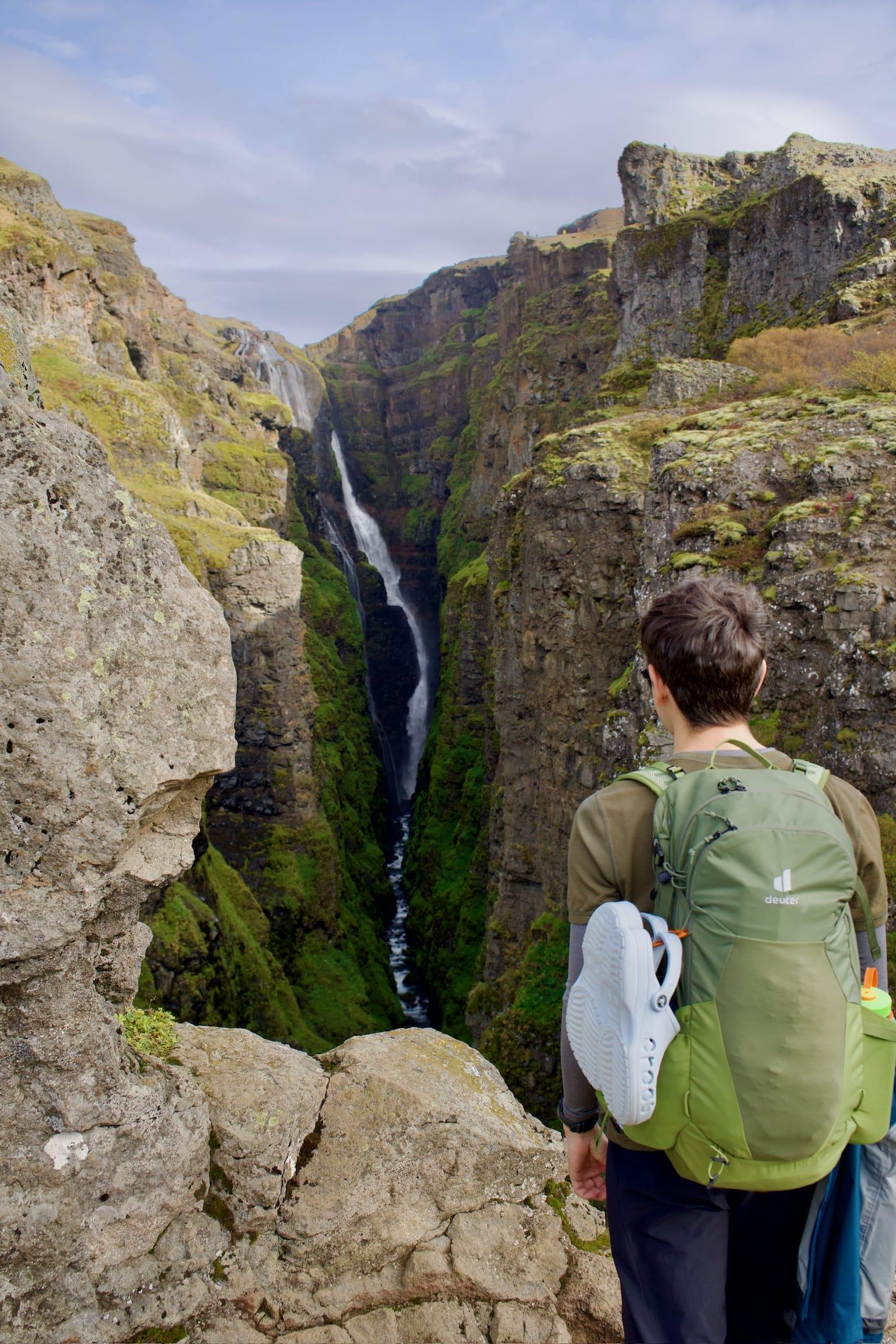 Ein Wanderer mit grünem Rucksack und Schuhen blickt auf eine dramatische Schlucht mit einem hohen, schmalen Wasserfall, der unter einem teilweise bewölkten Himmel steile, moosbewachsene Klippen hinabstürzt.