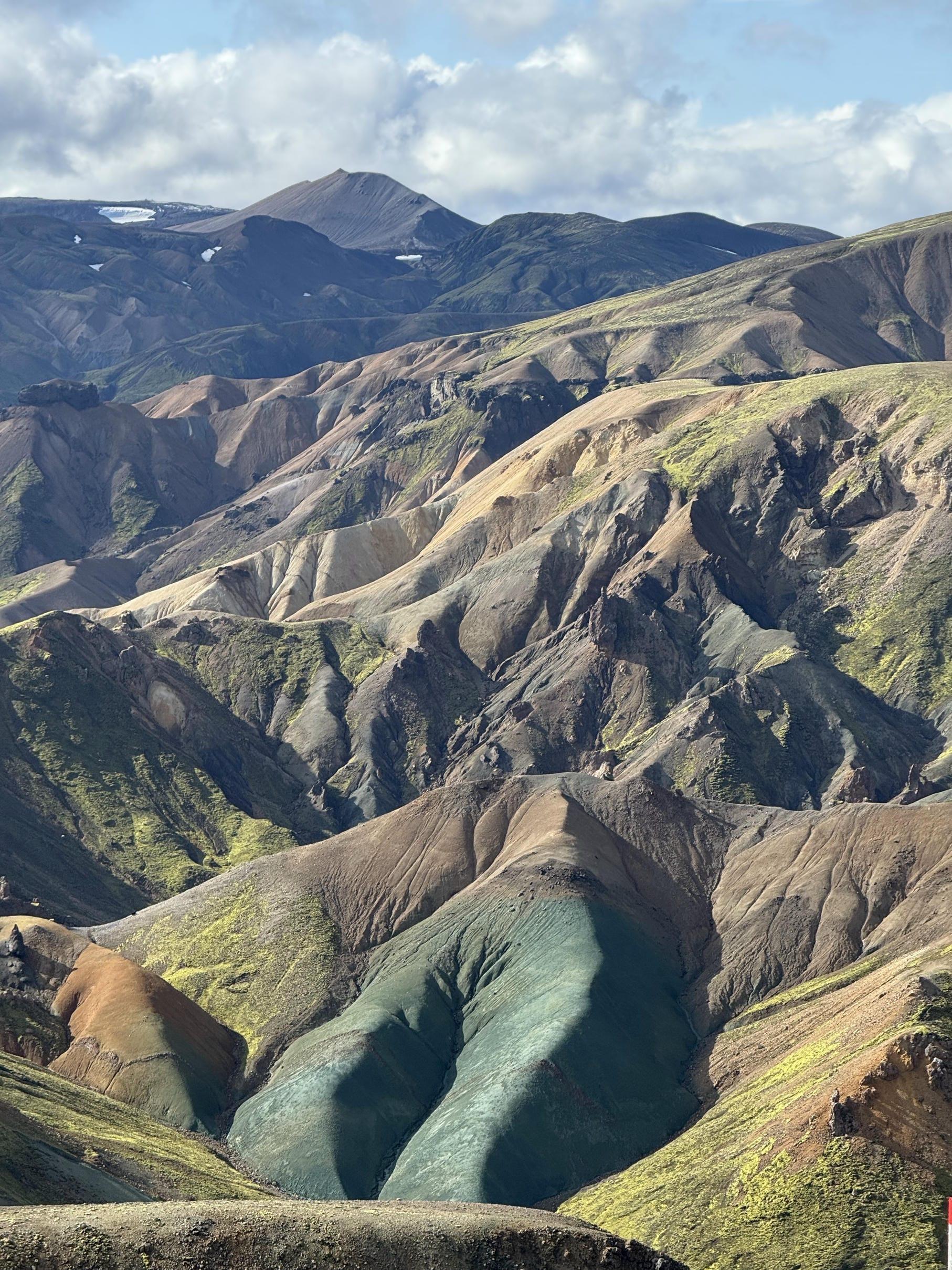 Farbenfrohe, zerklüftete Berge mit grünen, braunen und hellbraunen Gesteinsschichten, bedeckt mit Moosflecken unter einem teilweise bewölkten Himmel. Die Landschaft ist dramatisch und strukturiert, mit tiefen Tälern und Bergrücken.